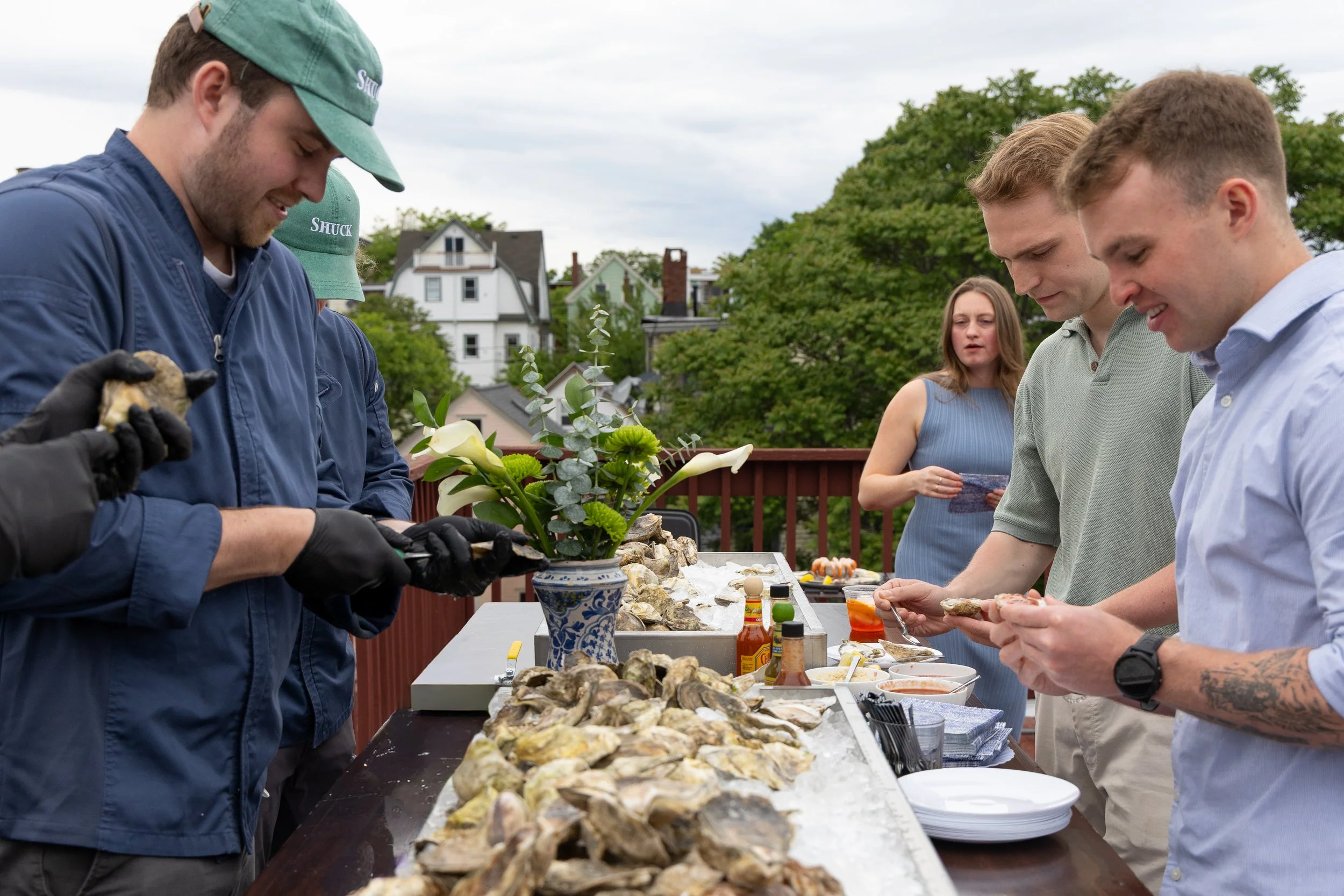 Group of people at an outdoor oyster shucking party, preparing oysters on a long table with condiments and a decorative vase with flowers, against a background of houses and green trees on a cloudy day. oyster bar boston new england