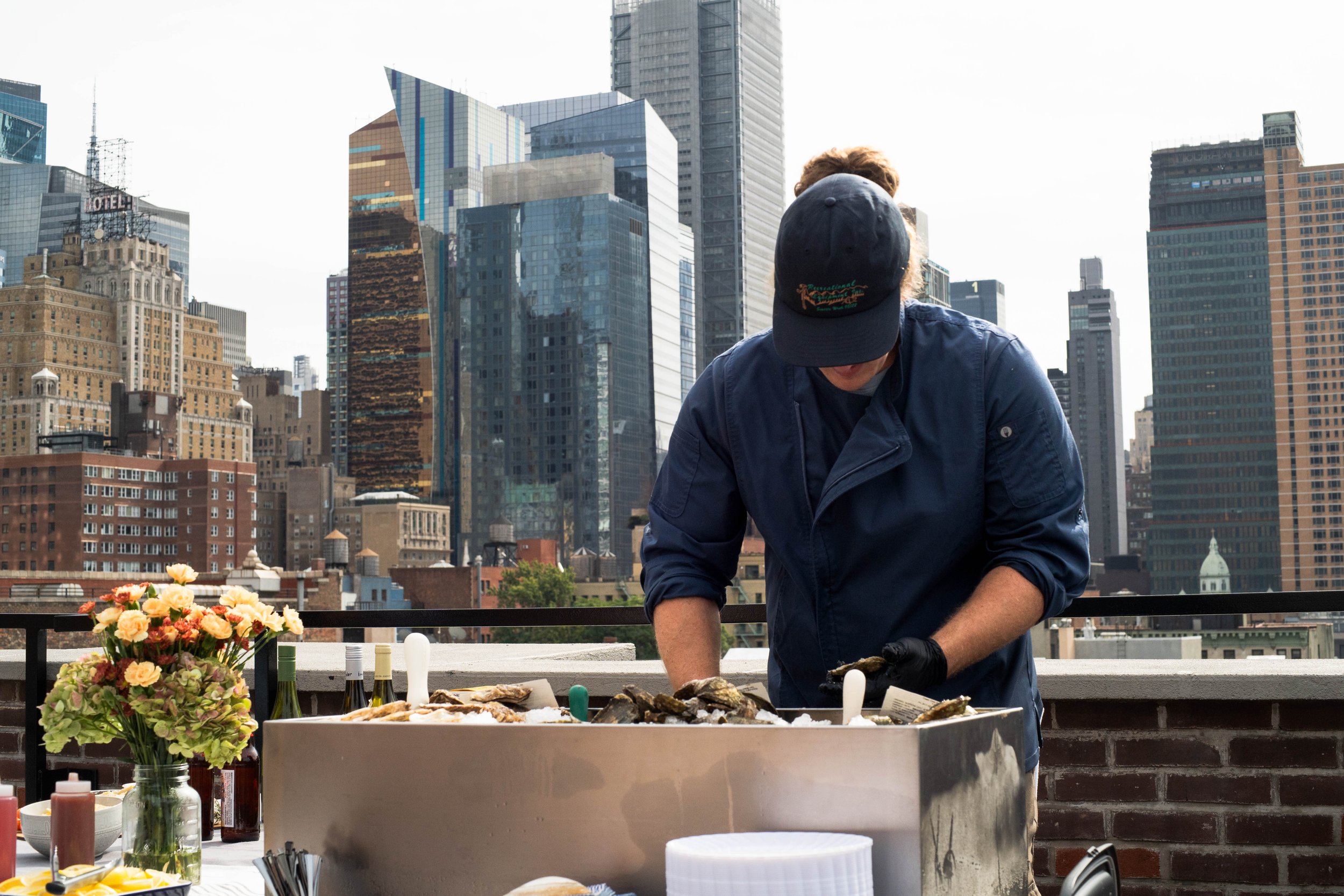 A man wearing a dark baseball cap with embroidered text, a chef jacker, and black gloves preparing seafood on a rooftop with a city skyline in the background. oyster bar boston new england