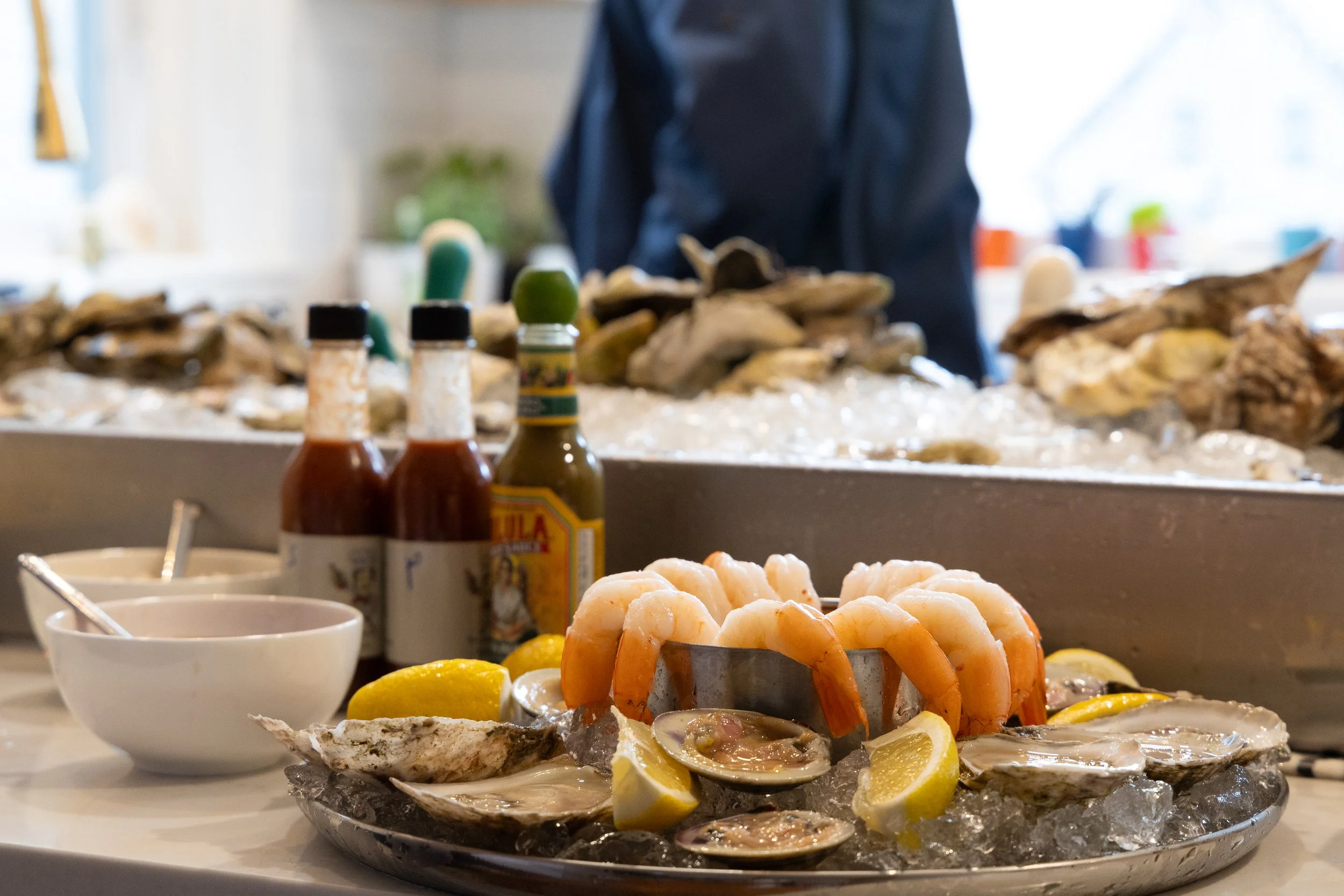 Seafood platter with oysters, shrimp, lemon wedges, and sauces on ice, with condiments and a person in the background. oyster bar boston new england