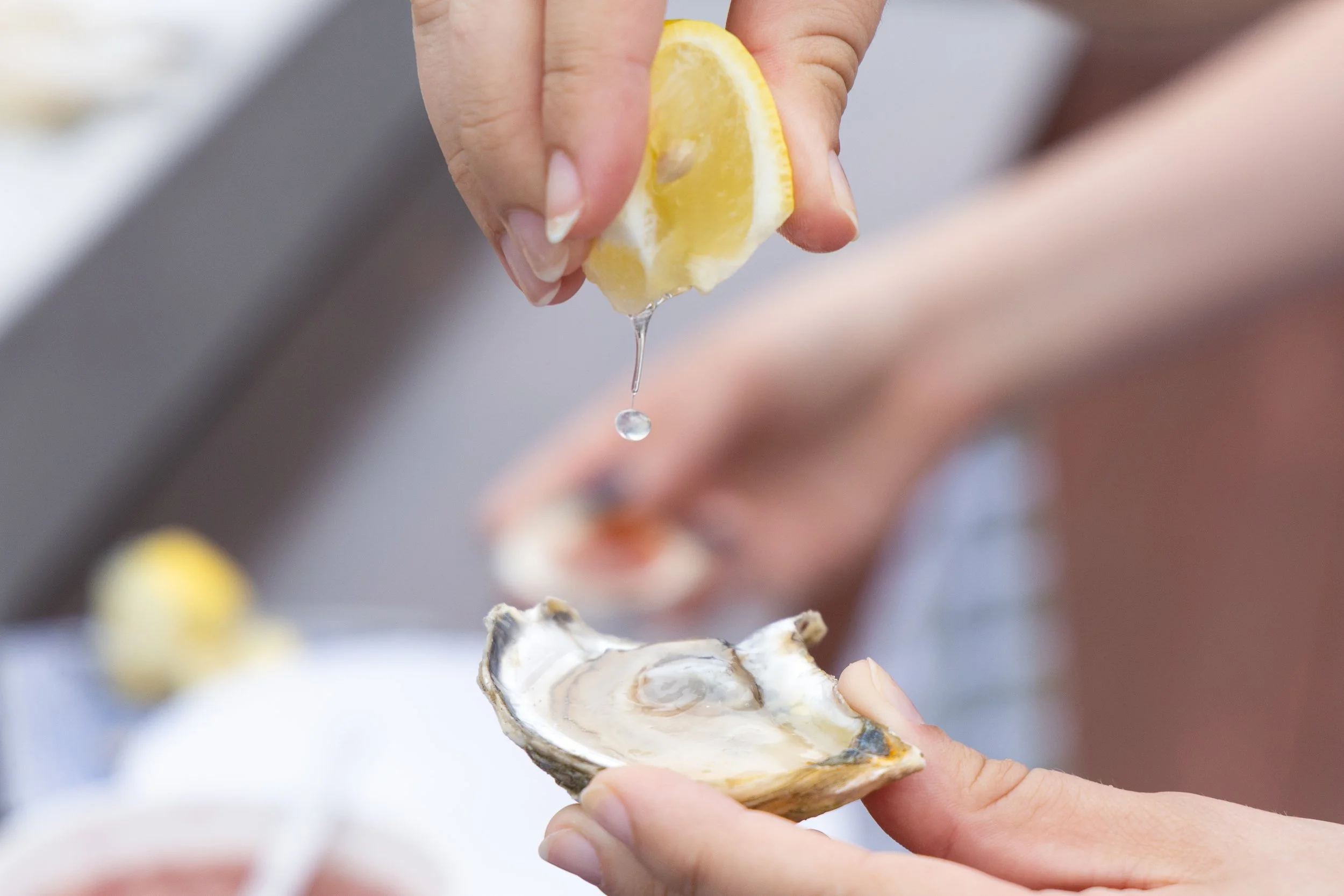 A person holding an open oyster shell with a lemon wedge above it, with water droplets falling from the lemon.