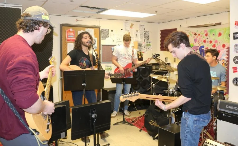 A group of five young musicians practicing in a small room with musical posters and equipment, including guitars and drums.