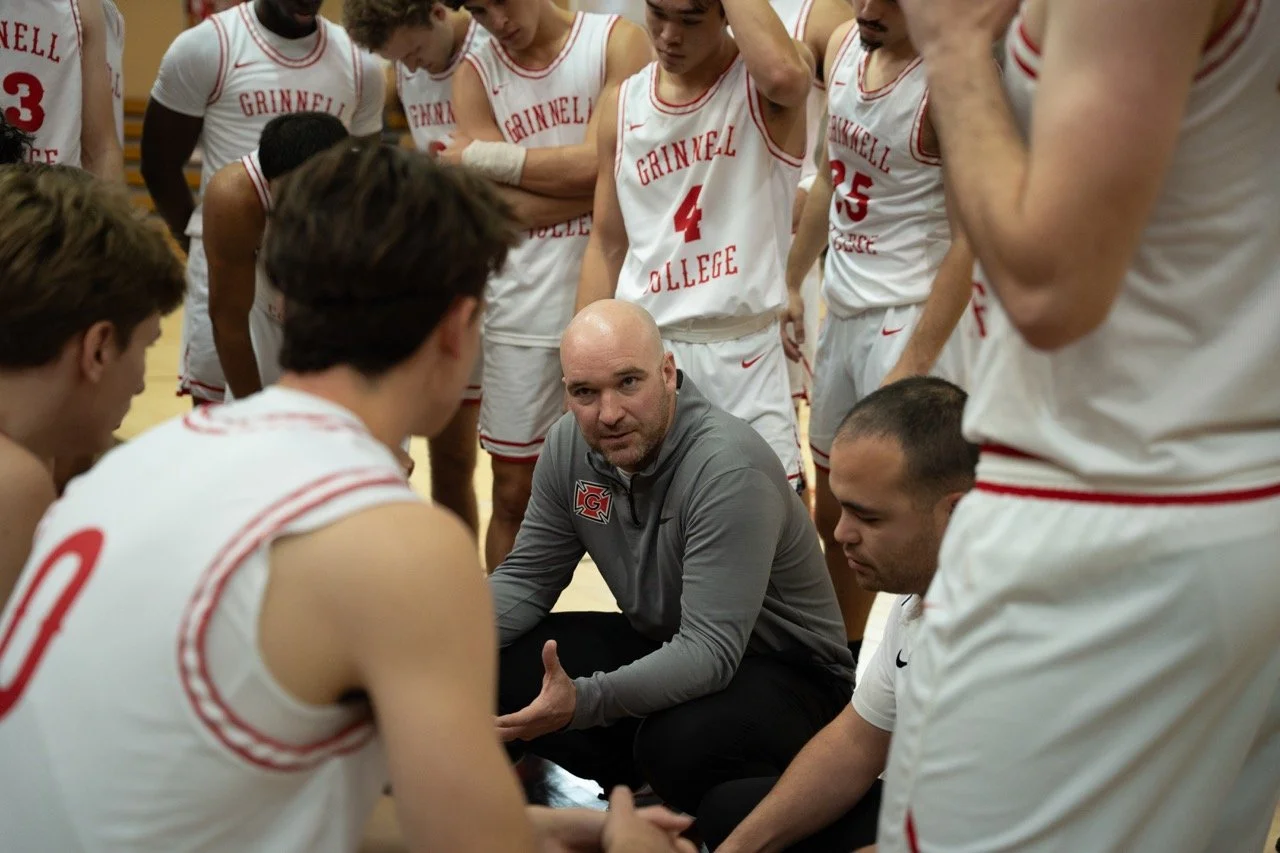 A coach giving a pep talk to a college basketball team during a timeout in a gymnasium.