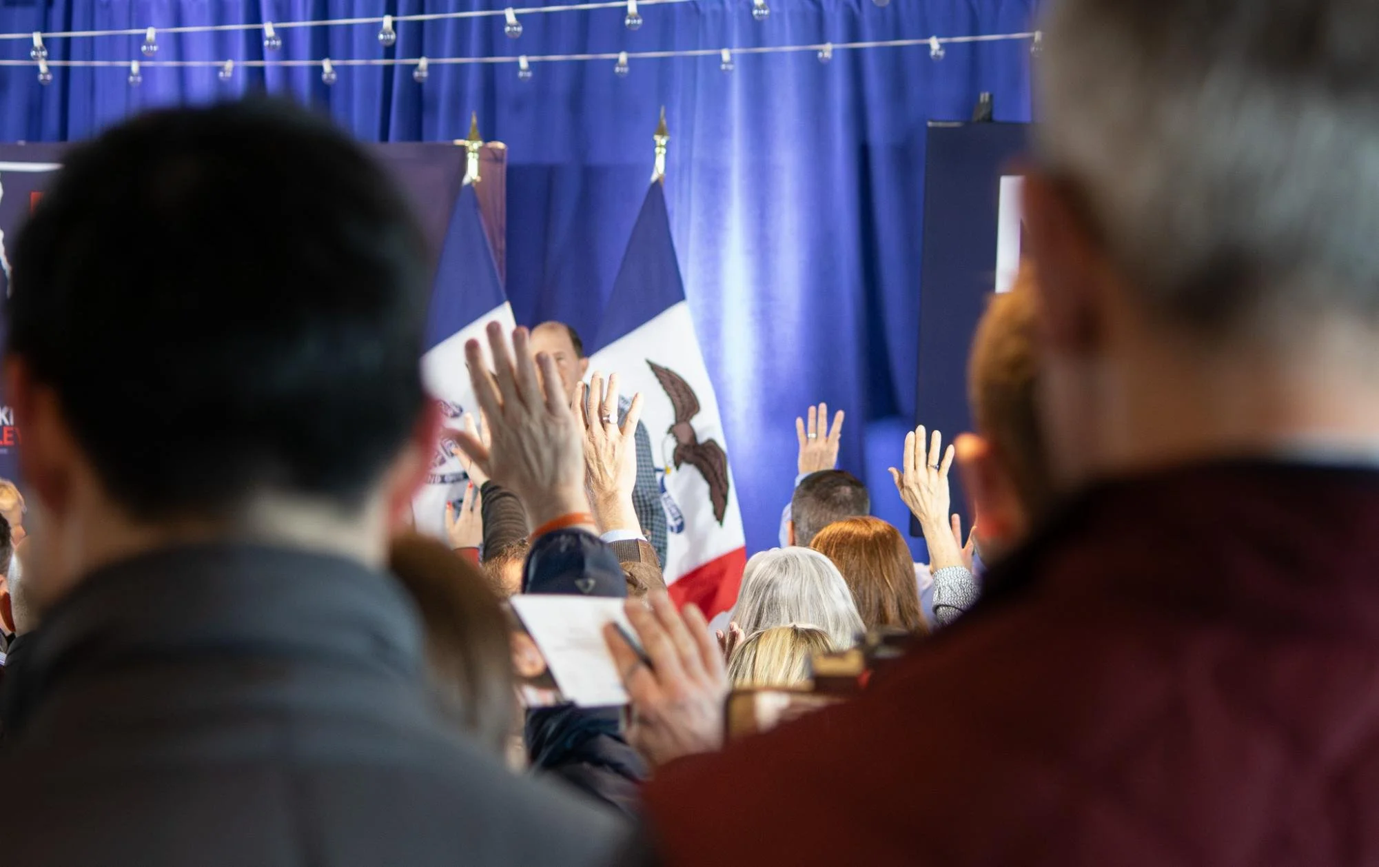 Audience at a political event with raised hands, facing a speaker in front of a blue curtain and Iowa state flags.