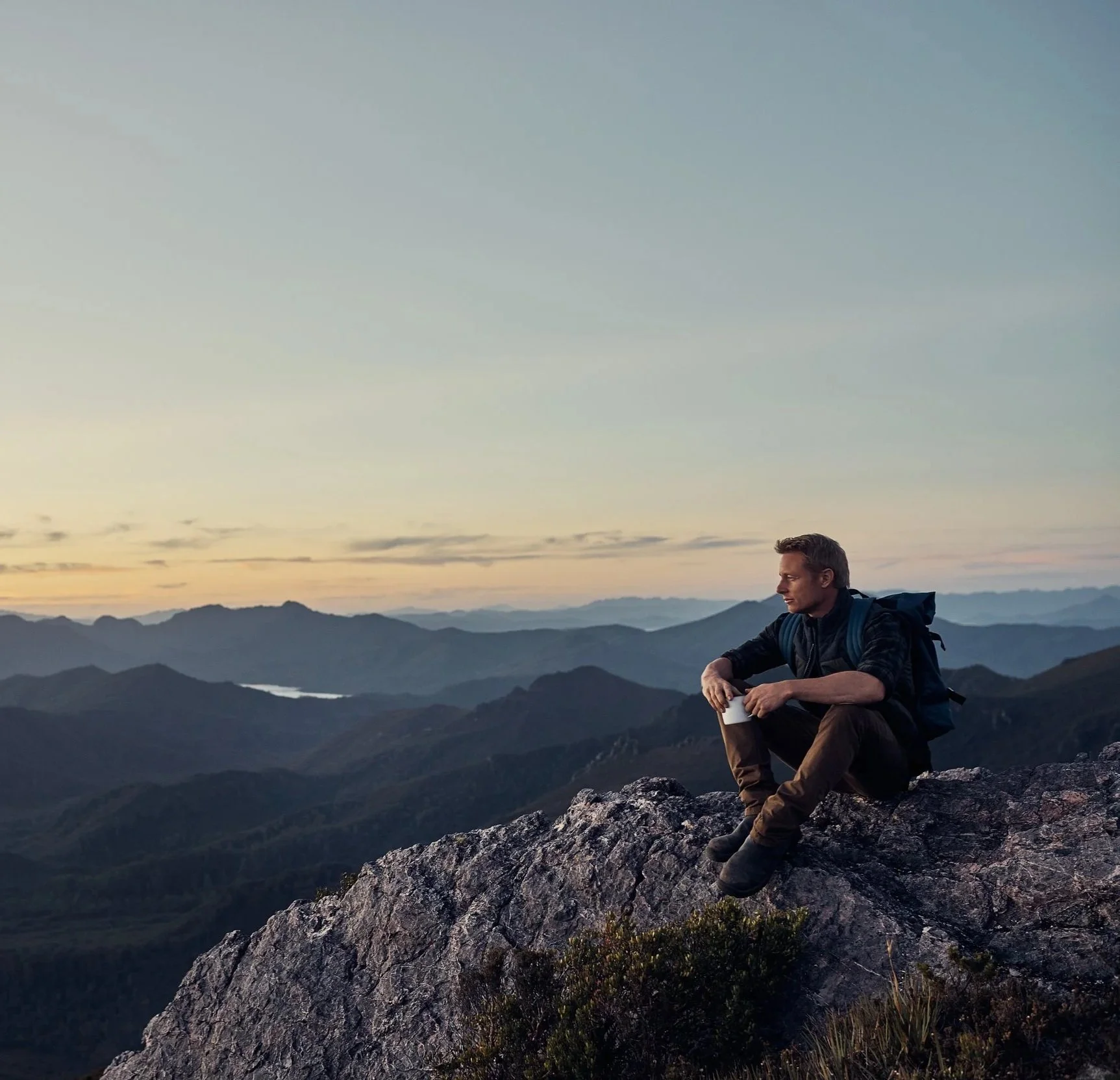 A man with a backpack sitting on a mountain rock, looking contemplatively at the distant mountain ranges at sunset.