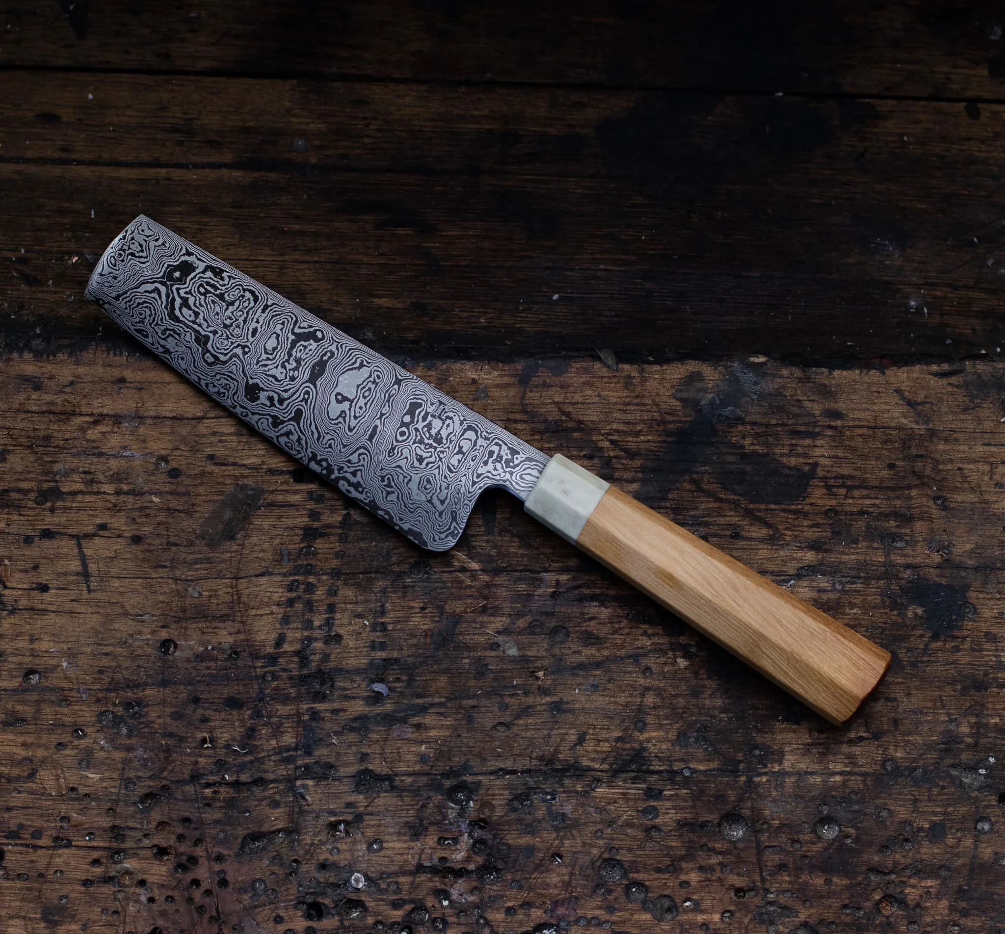 A Damascus knife with a patterned blade and a wooden handle resting on a dark, distressed wooden surface.