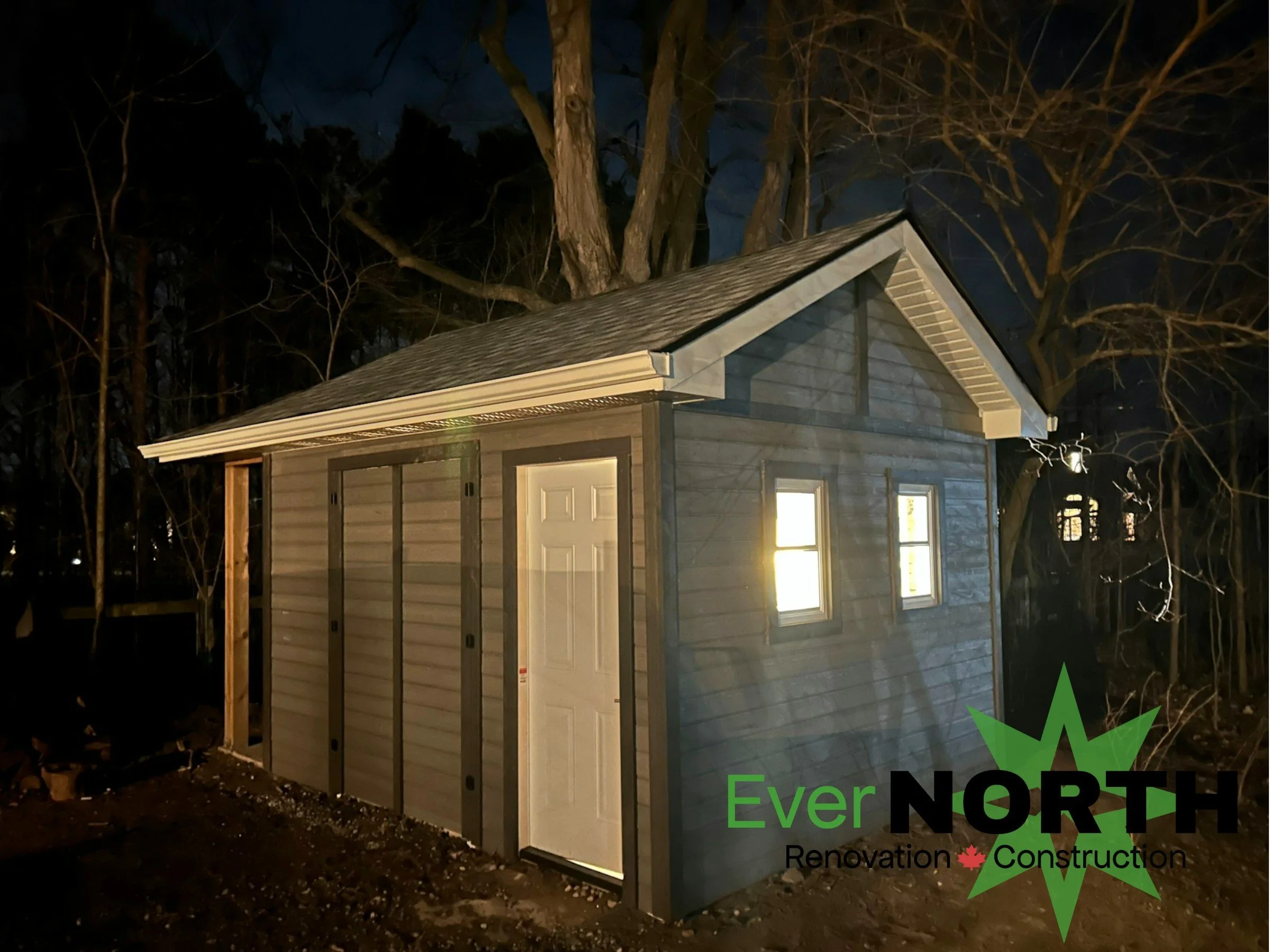 A small shed at night with illuminated windows, gray siding, a gable roof, and a white door. There are trees in the background and a logo in the bottom right corner for Ever North Renovation & Construction.