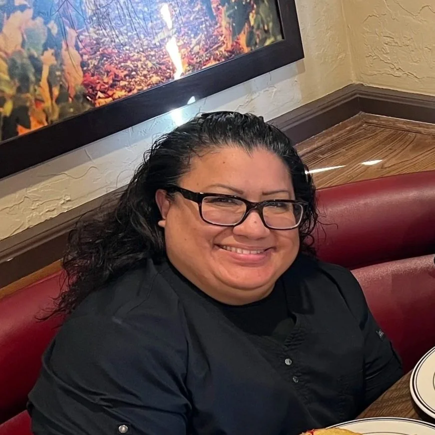 A woman with dark curly hair, glasses, and a black shirt, smiling while sitting at a table in a restaurant with red seating and a colorful landscape painting on the wall behind her.