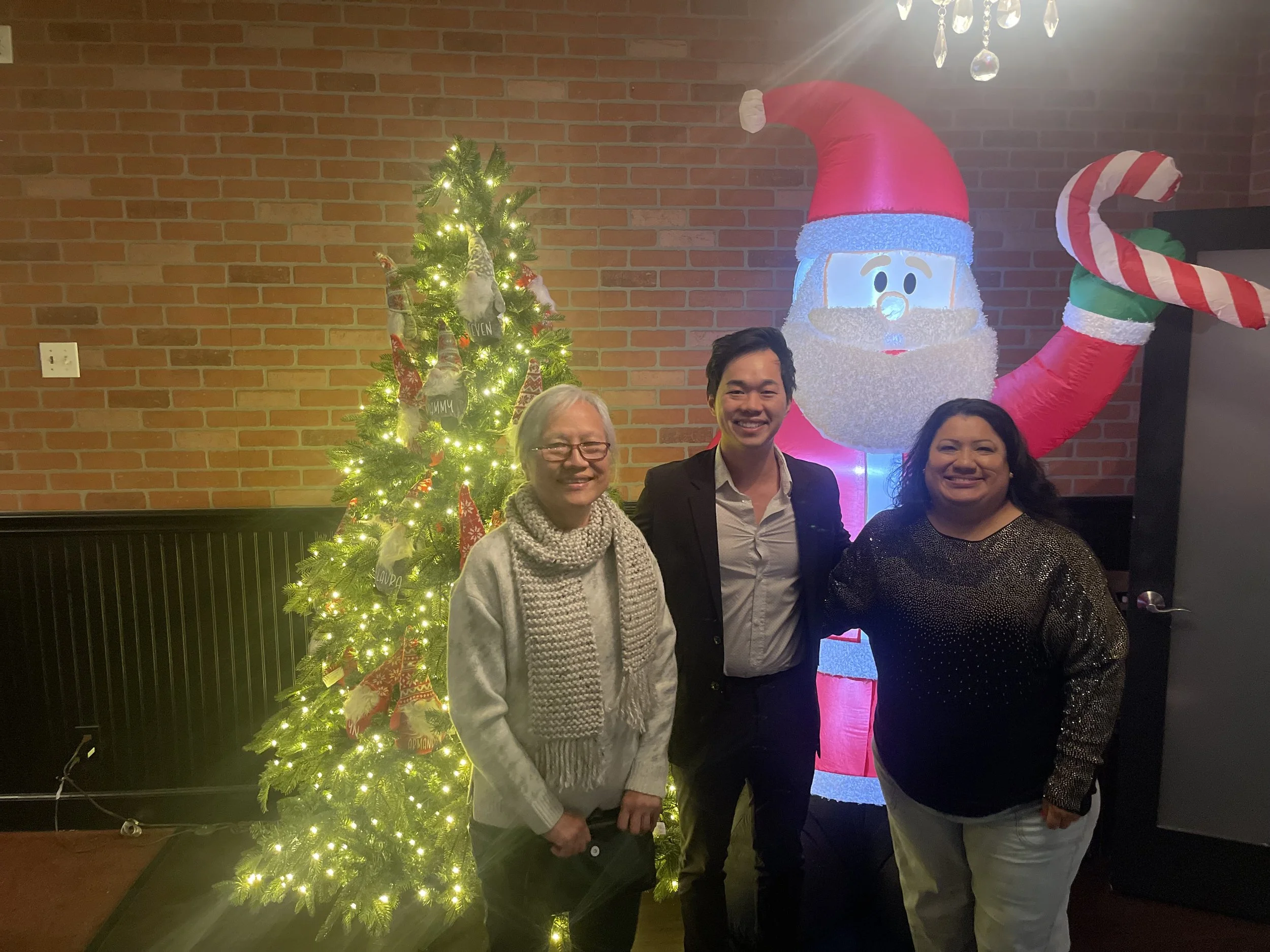 Three people standing in front of a decorated Christmas tree and an inflatable Santa Claus with a candy cane arm, smiling at the camera.