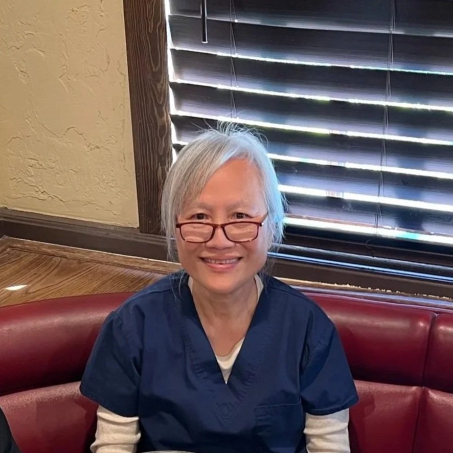 An elderly woman with short gray hair, glasses, and a bright smile is sitting on a red leather booth in a restaurant with striped blinds and textured beige walls.