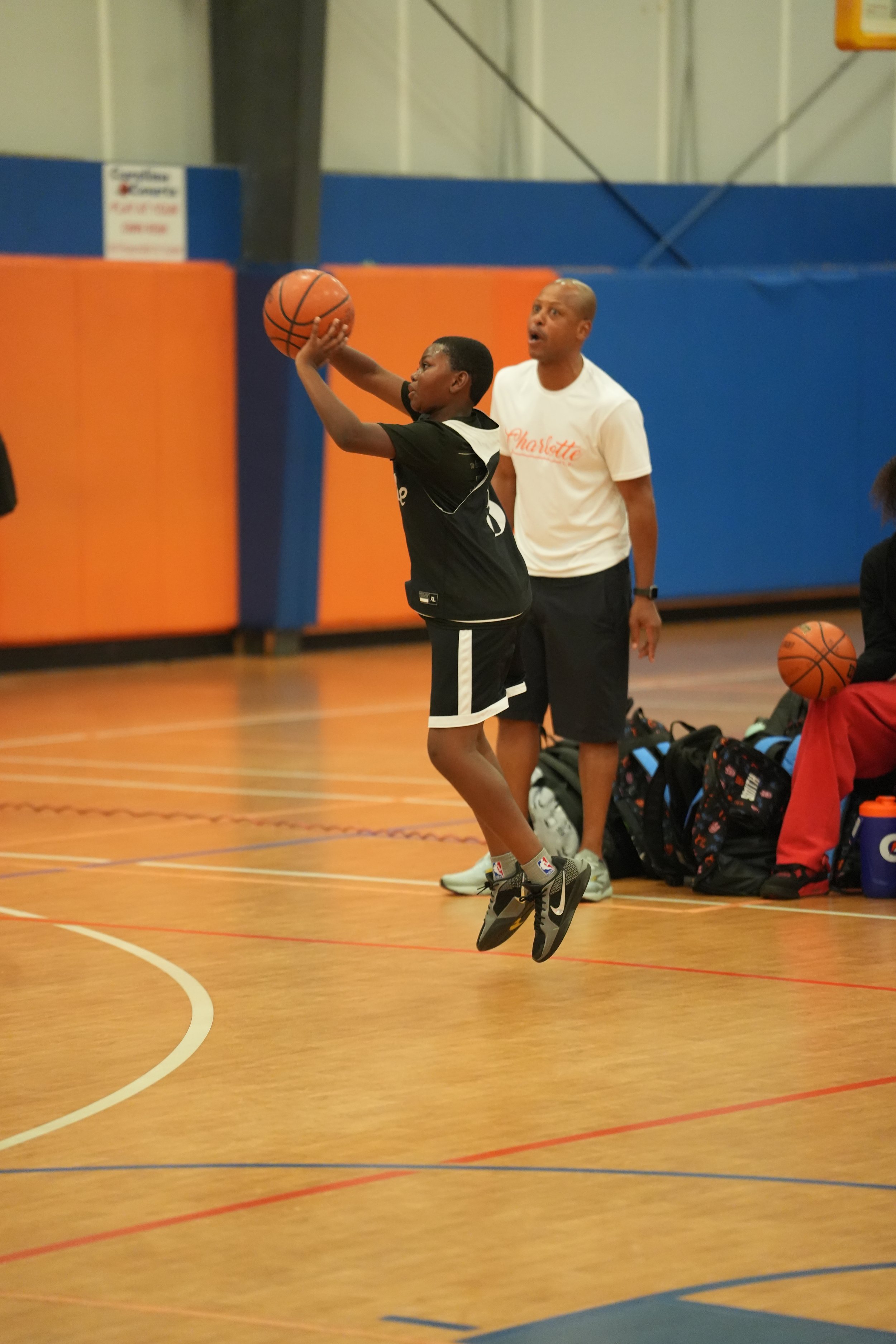 A young boy jumps in the air while holding a basketball, preparing to take a shot during a basketball practice in an indoor gym. An adult coach and others are visible in the background.