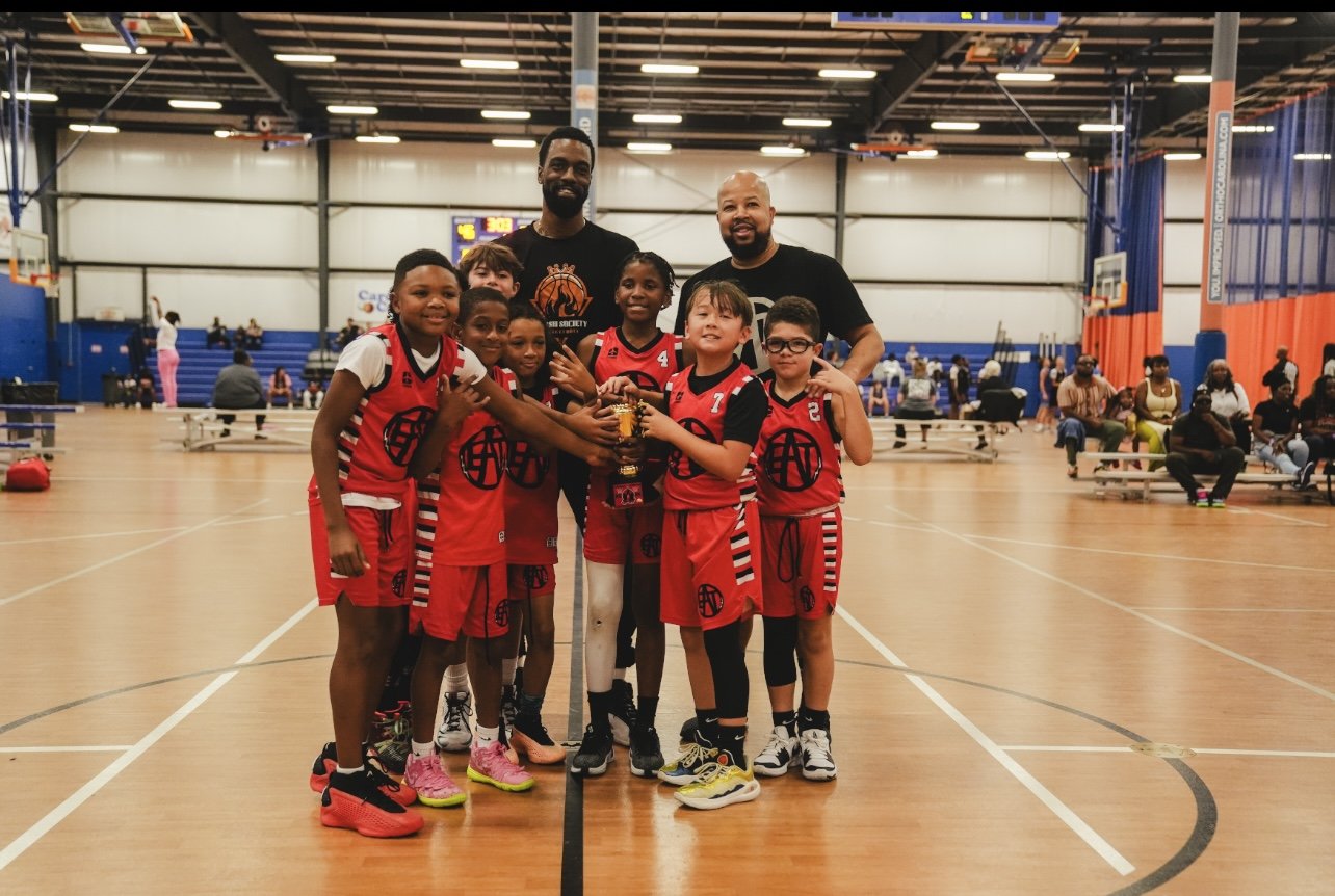 Young girls' basketball team in red uniforms celebrating with two coaches holding a trophy in an indoor gymnasium.