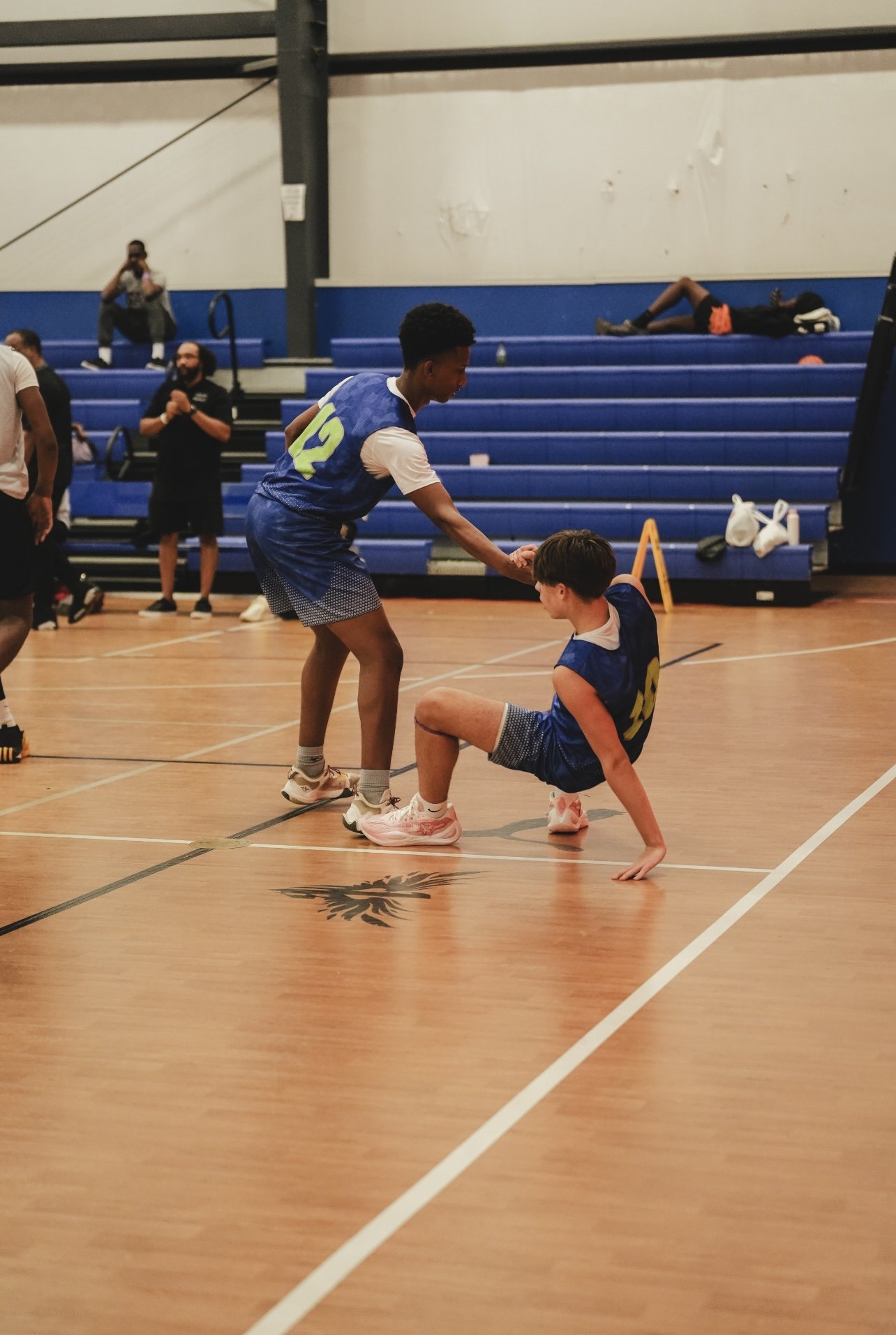 Two young basketball players, wearing blue jerseys, are on an indoor court. One is standing and holding the arm of the other, who is sitting on the floor with one hand on the ground. In the background, other people watch and sit on blue bleachers.