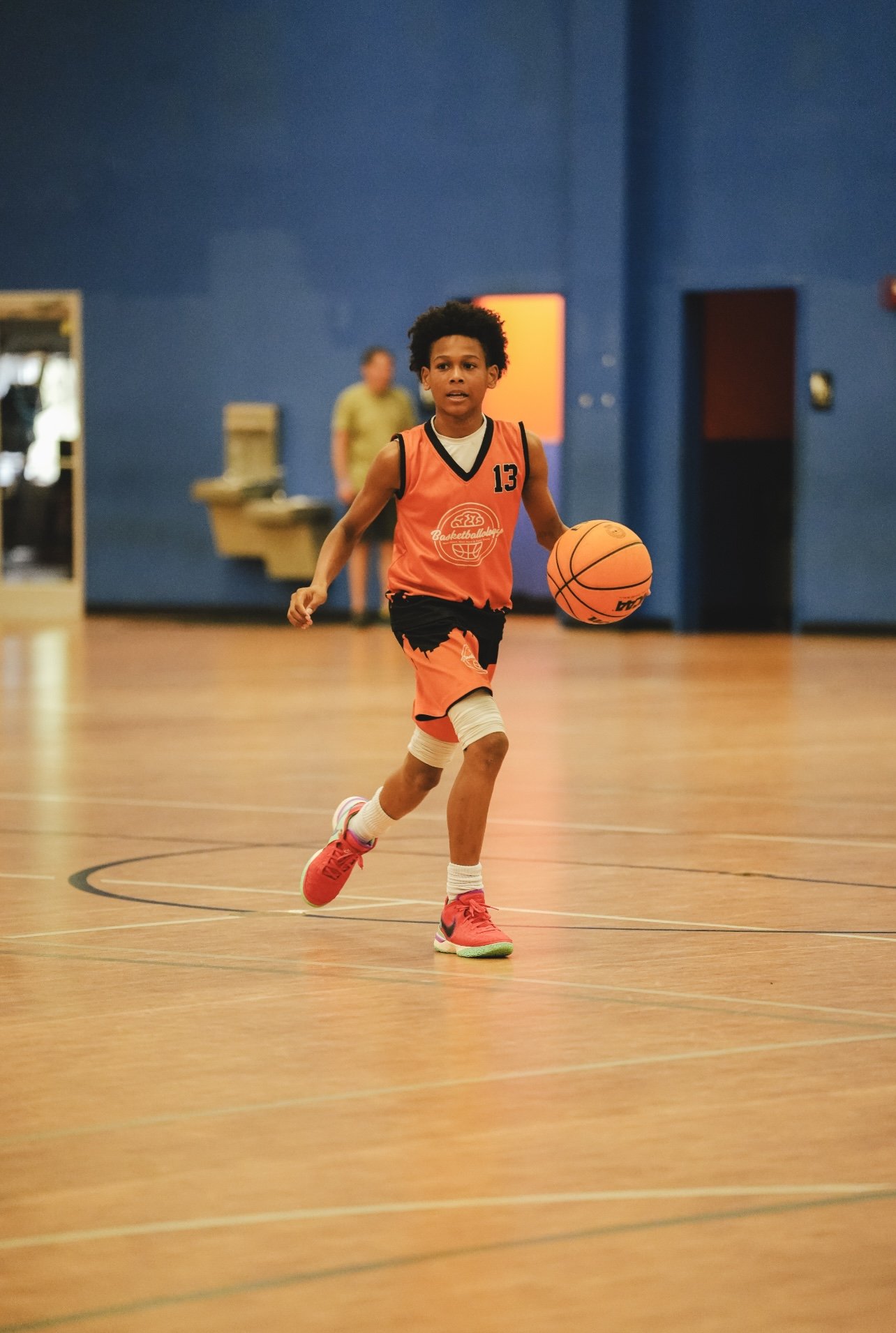 Young boy playing basketball indoors on a wooden court, wearing an orange sports uniform with the number 13.