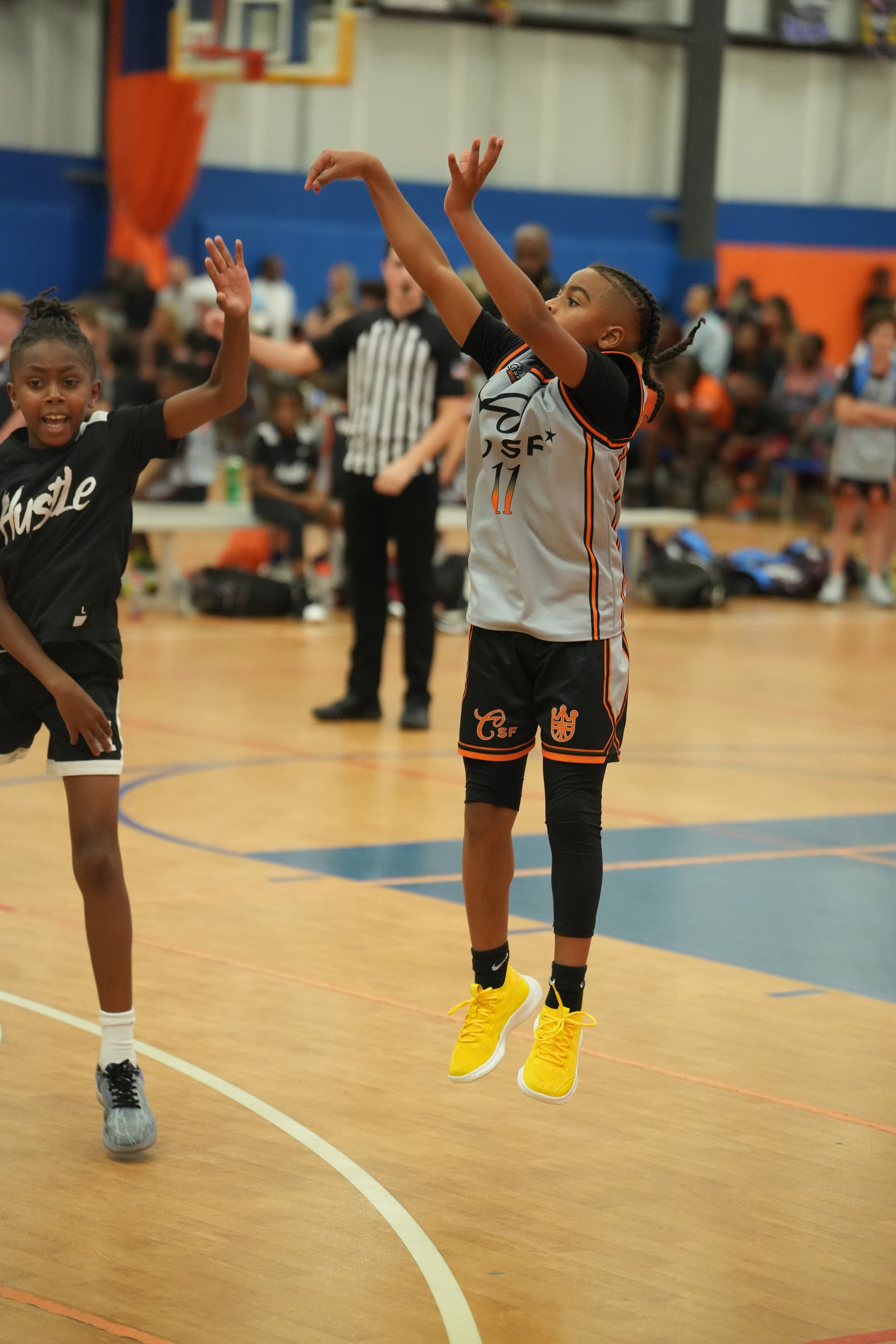 A young girl in a black sports jersey with the word 'Hustle' is playing basketball on an indoor court, jumping with her arm raised, as another girl in a white and black uniform with yellow shoes attempts a shot or pass, with a crowd of spectators in