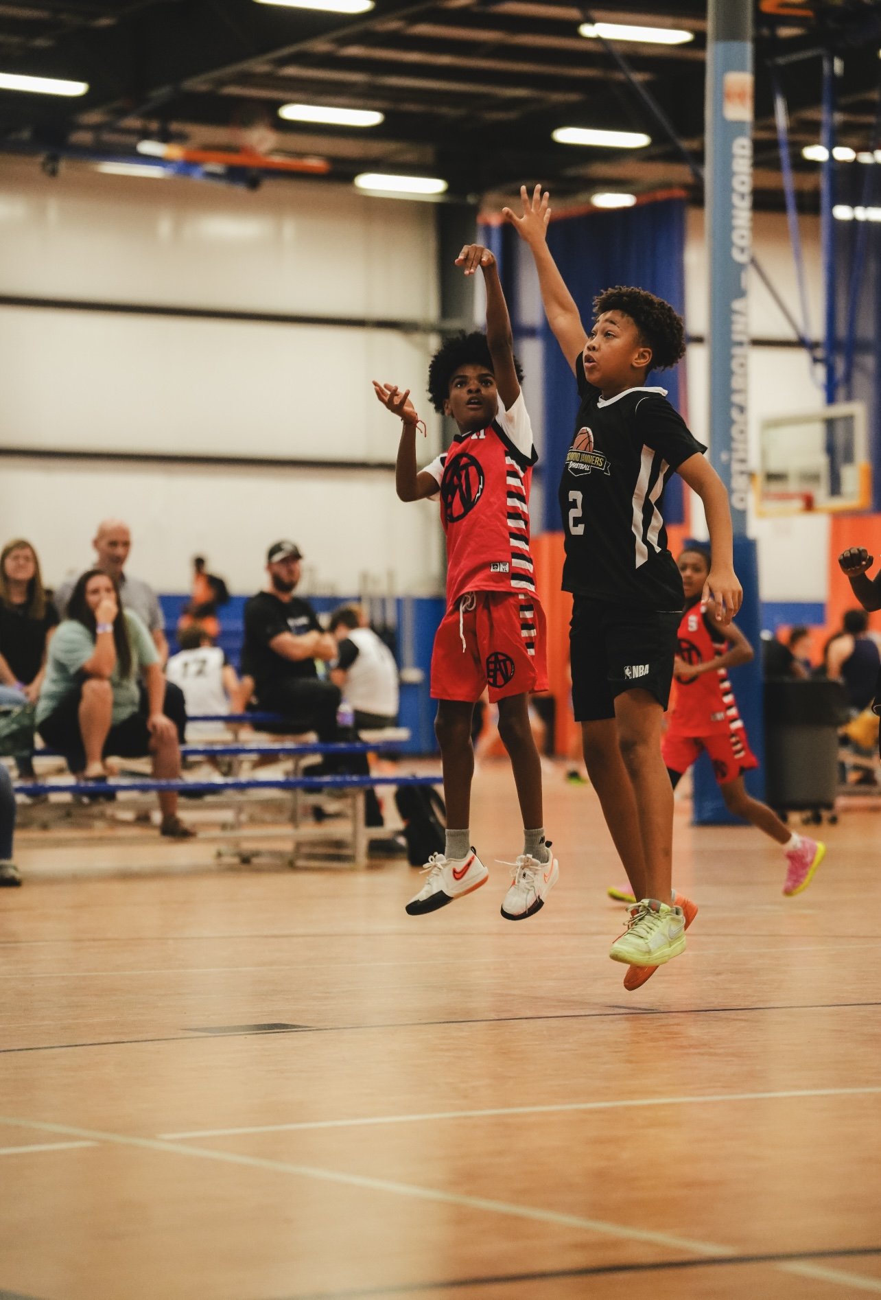 Two young boys playing basketball and jumping during a game in an indoor gym, with spectators watching in the background.