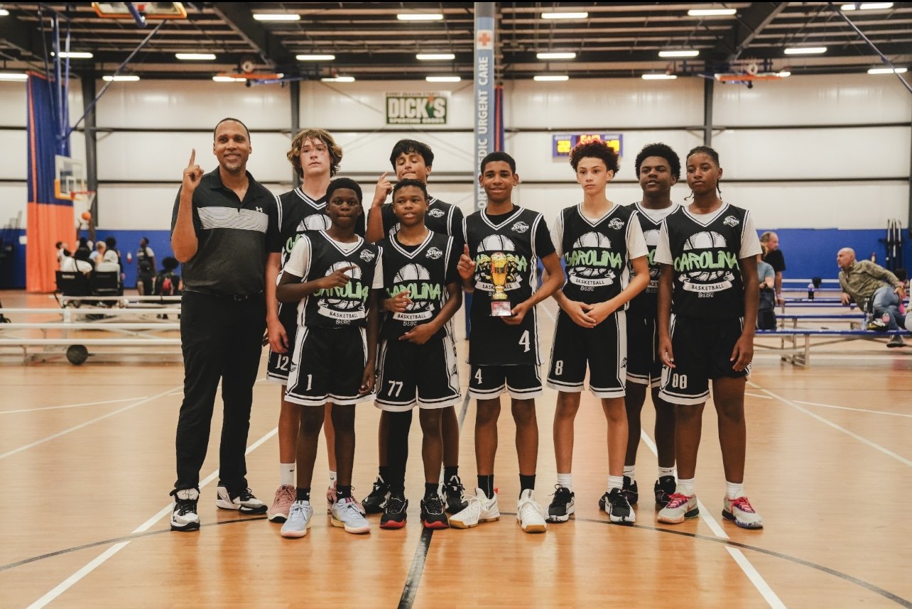 A youth basketball team in black and white uniforms posing with their coach in an indoor gym, with one player holding a trophy.