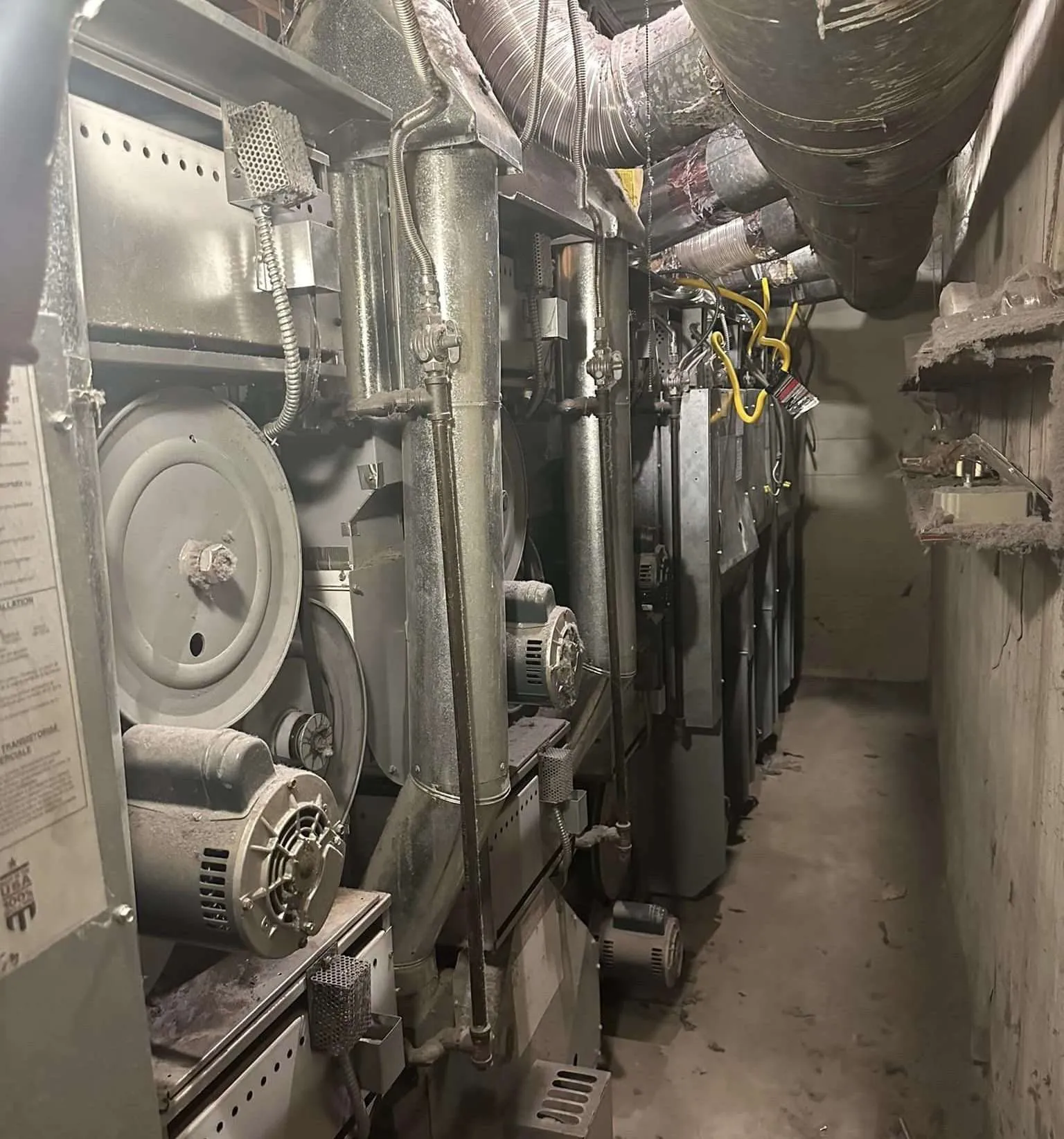 A narrow utility room with industrial HVAC or ventilation equipment and ductwork along the ceiling.