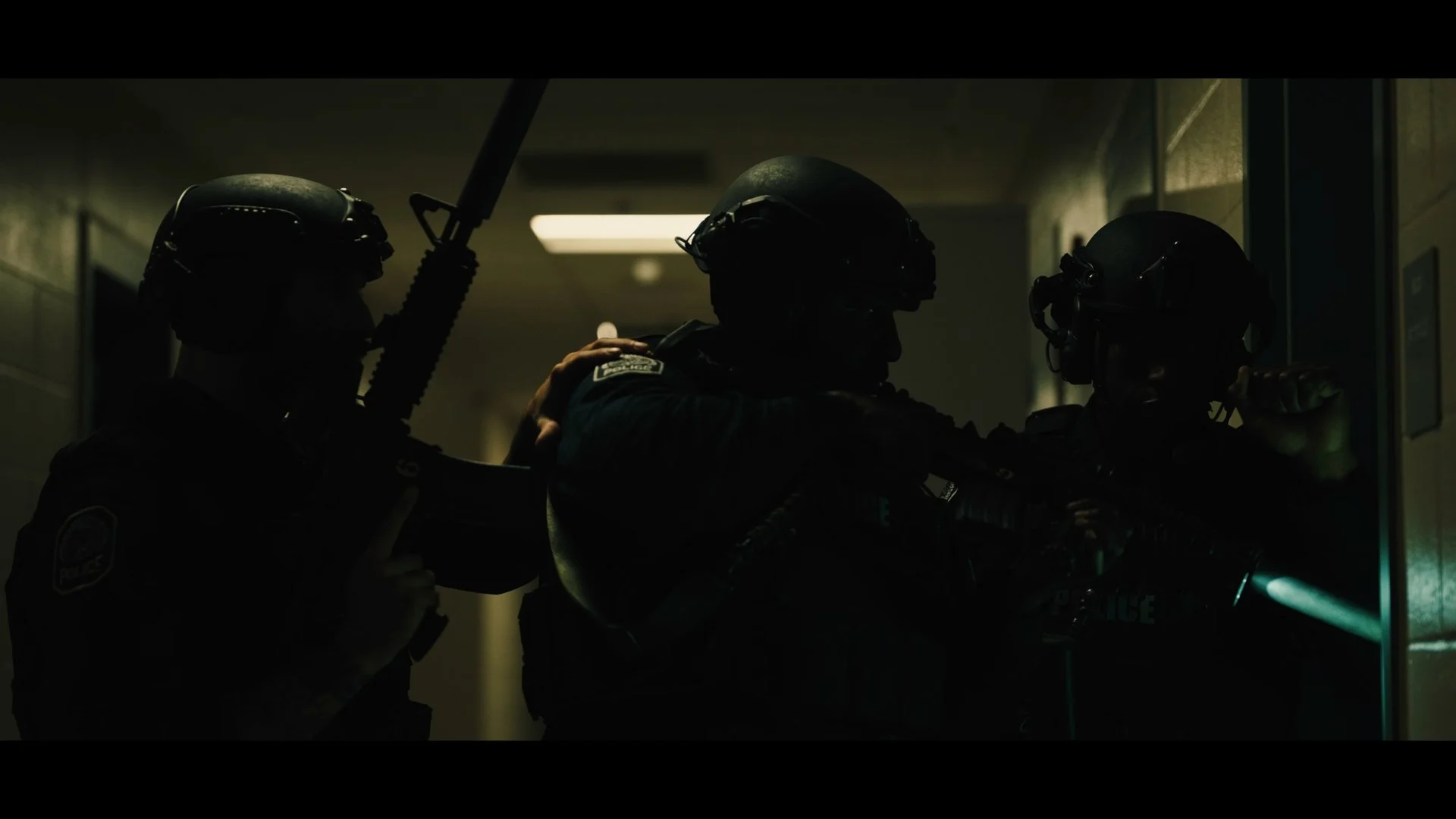 Dark silhouette of police officers in tactical gear inside a hallway, holding a shield and a rifle, with subdued lighting and a faint light source in the background.