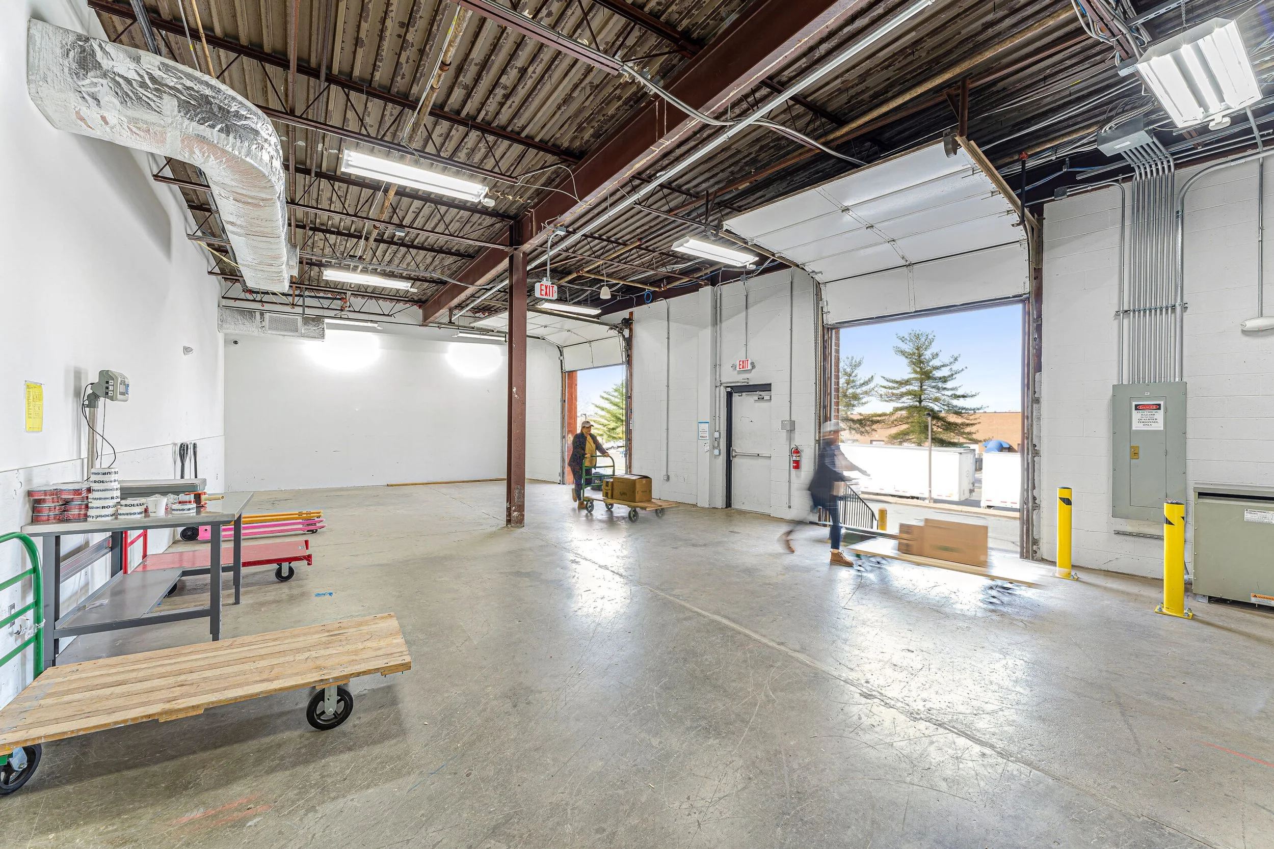 Interior of a warehouse or loading dock area with open garage doors, workers moving boxes with carts, and equipment along the walls.