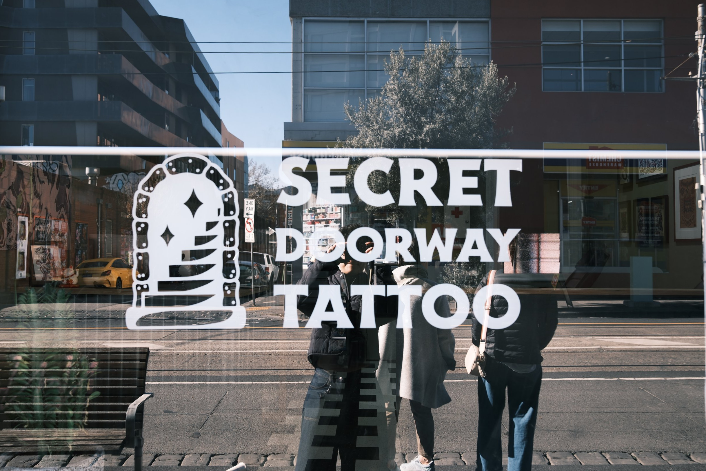 Glass window with white lettering stating 'Secret Doorway Tattoo' and a logo of a door with a diamond-shaped window. Reflections of people, parked cars, buildings, and street scene are visible in the glass.
