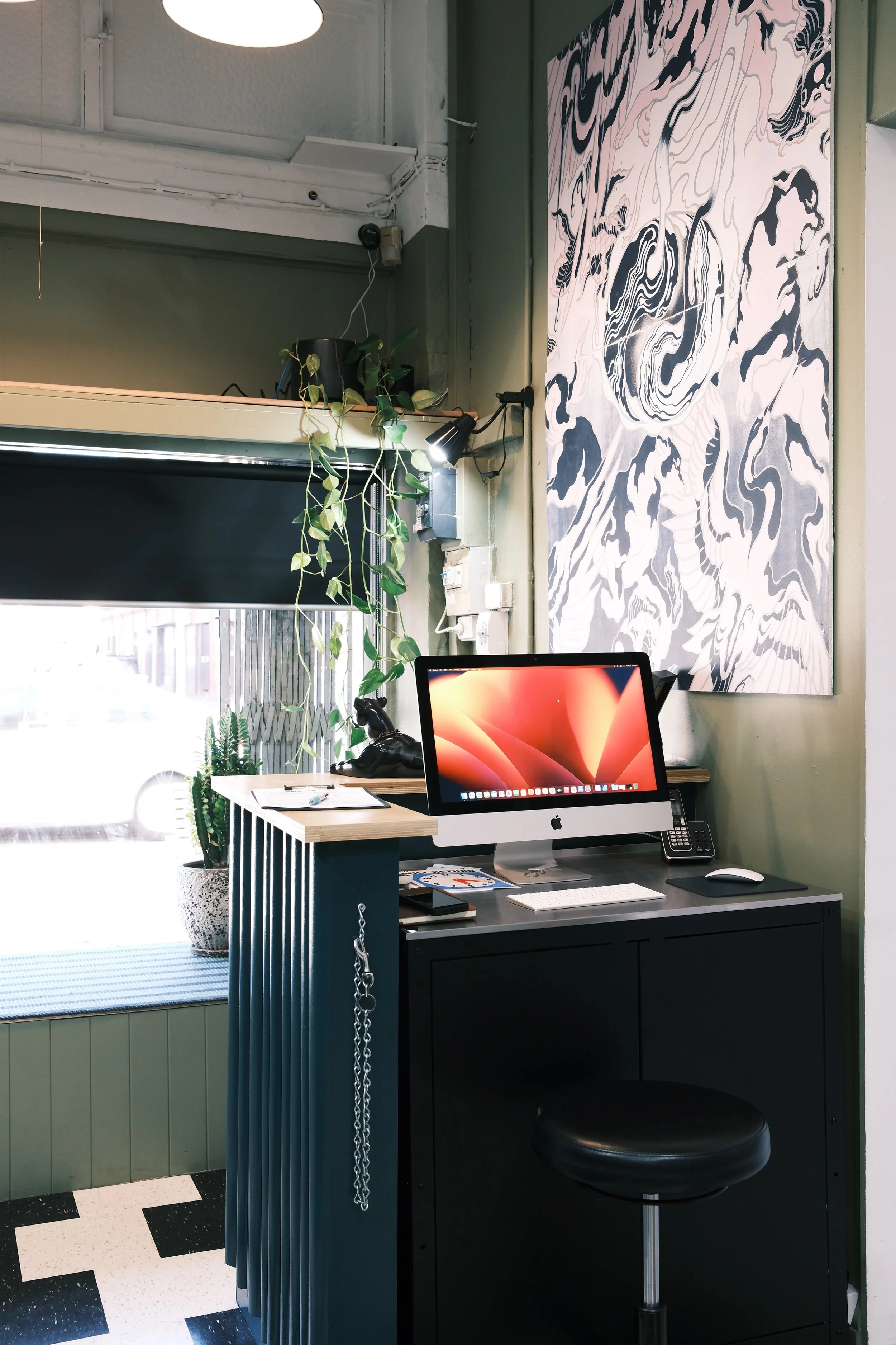 Office workspace with a desktop computer, a black and white abstract art piece on the wall, and green plants near a window.