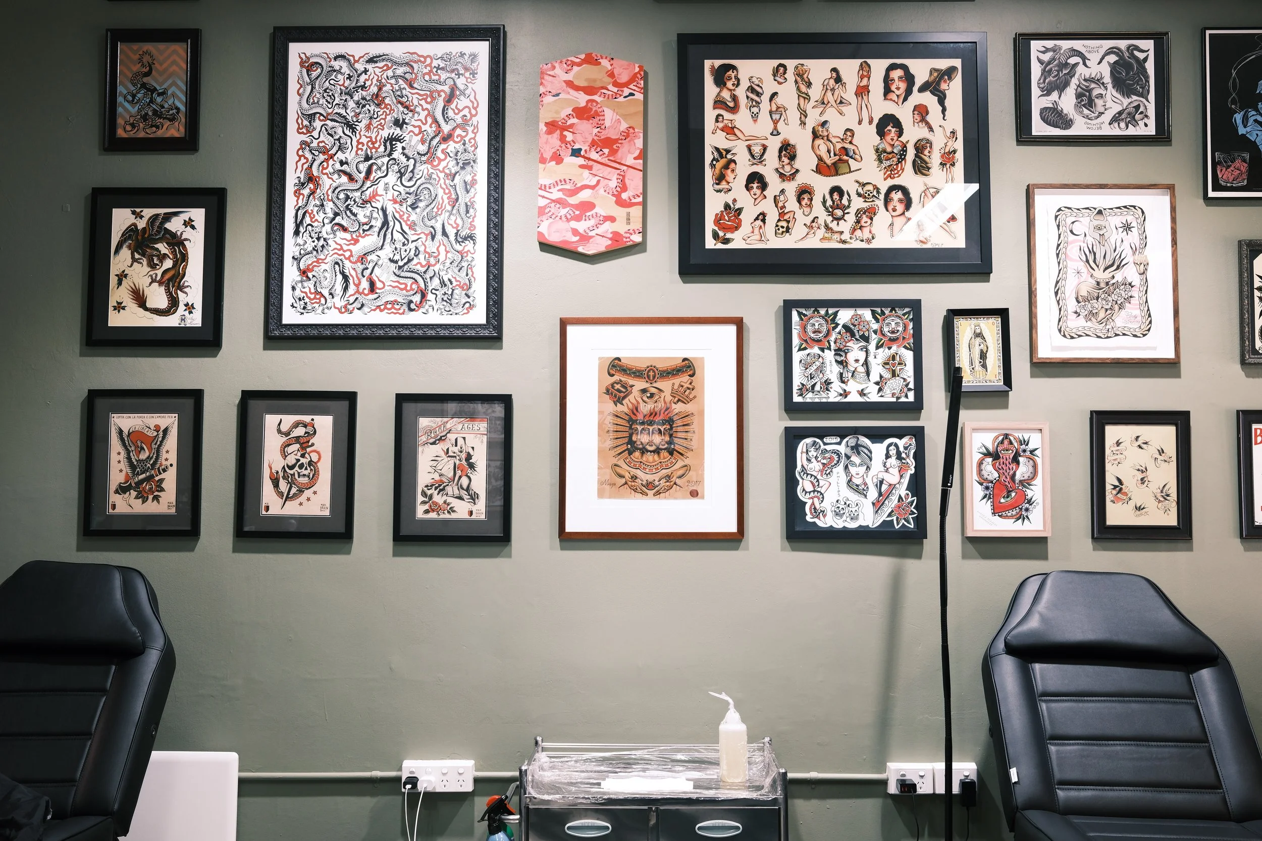 Wall decorated with various framed artwork, including traditional tattoo designs, dragon images, and portraits, in a room with black chairs and a hand sanitizer dispenser.
