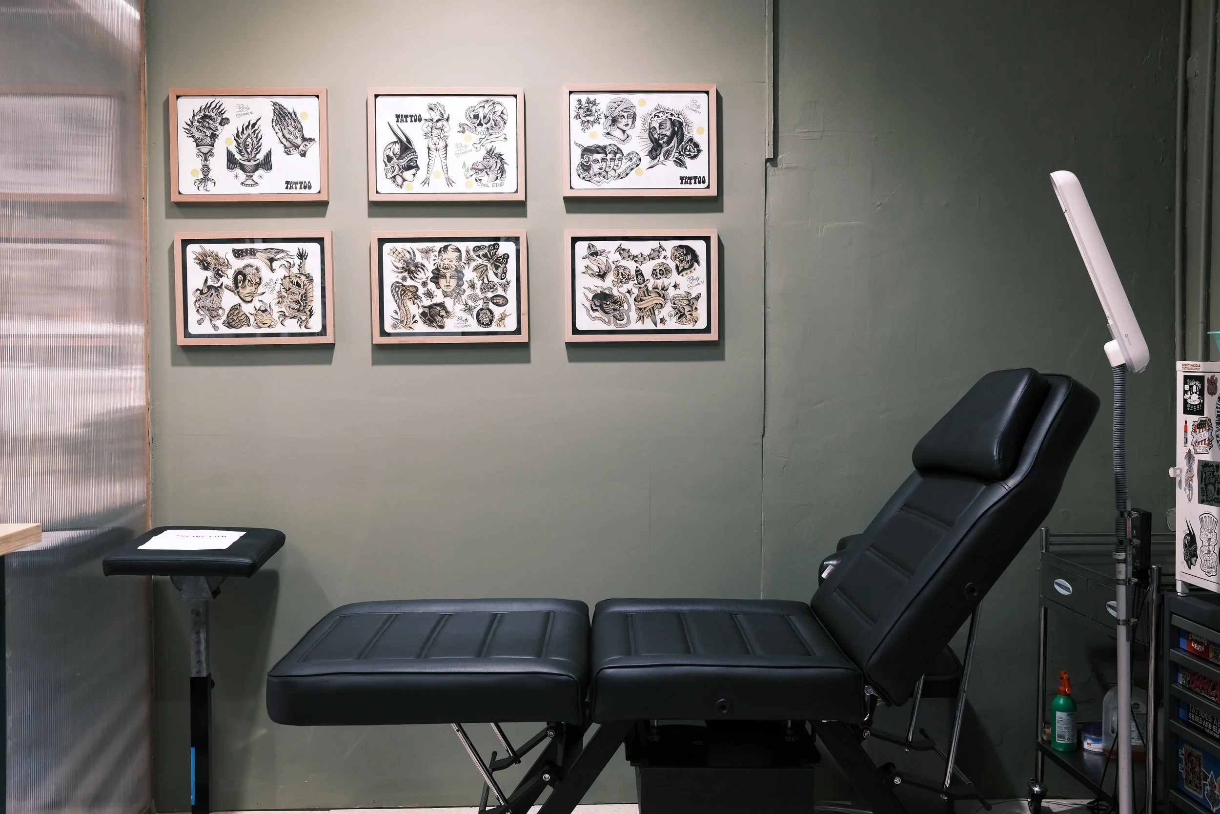 Tattoo parlor room with framed tattoo art on green wall, black reclining chair, and tattoo equipment.