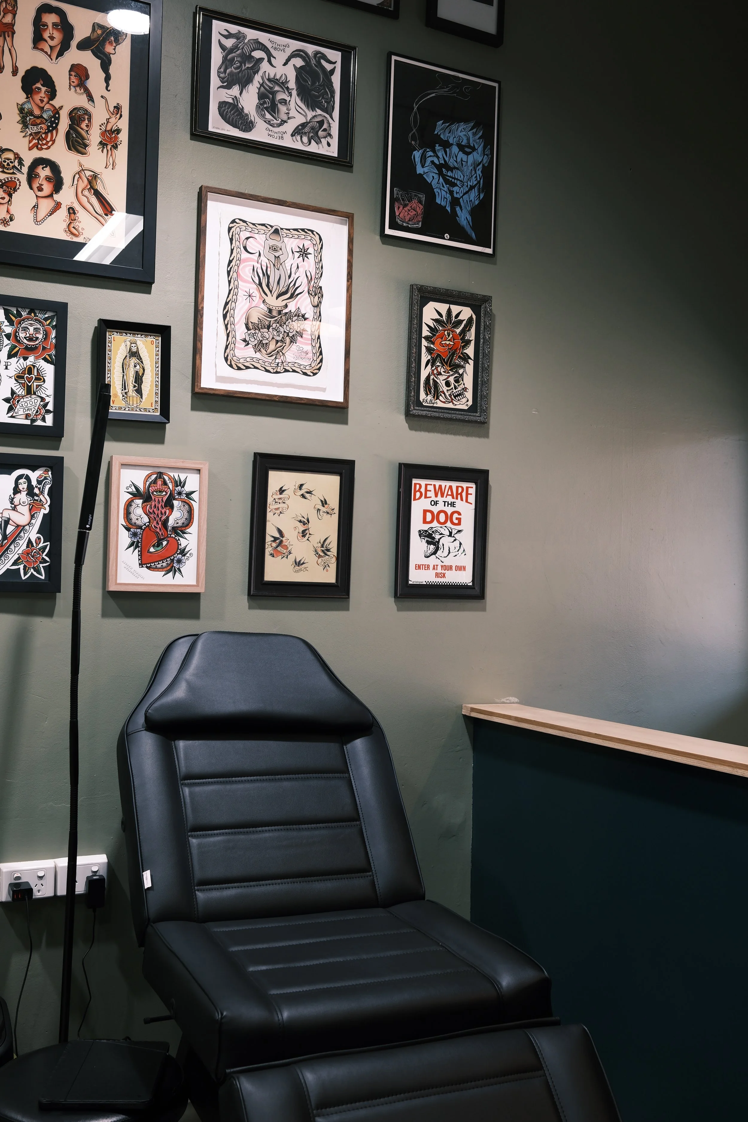 A tattoo studio waiting area with a black leather chair, wall art including framed tattoo designs, a 'Beware of the Dog' sign, and a tattoo machine stand.