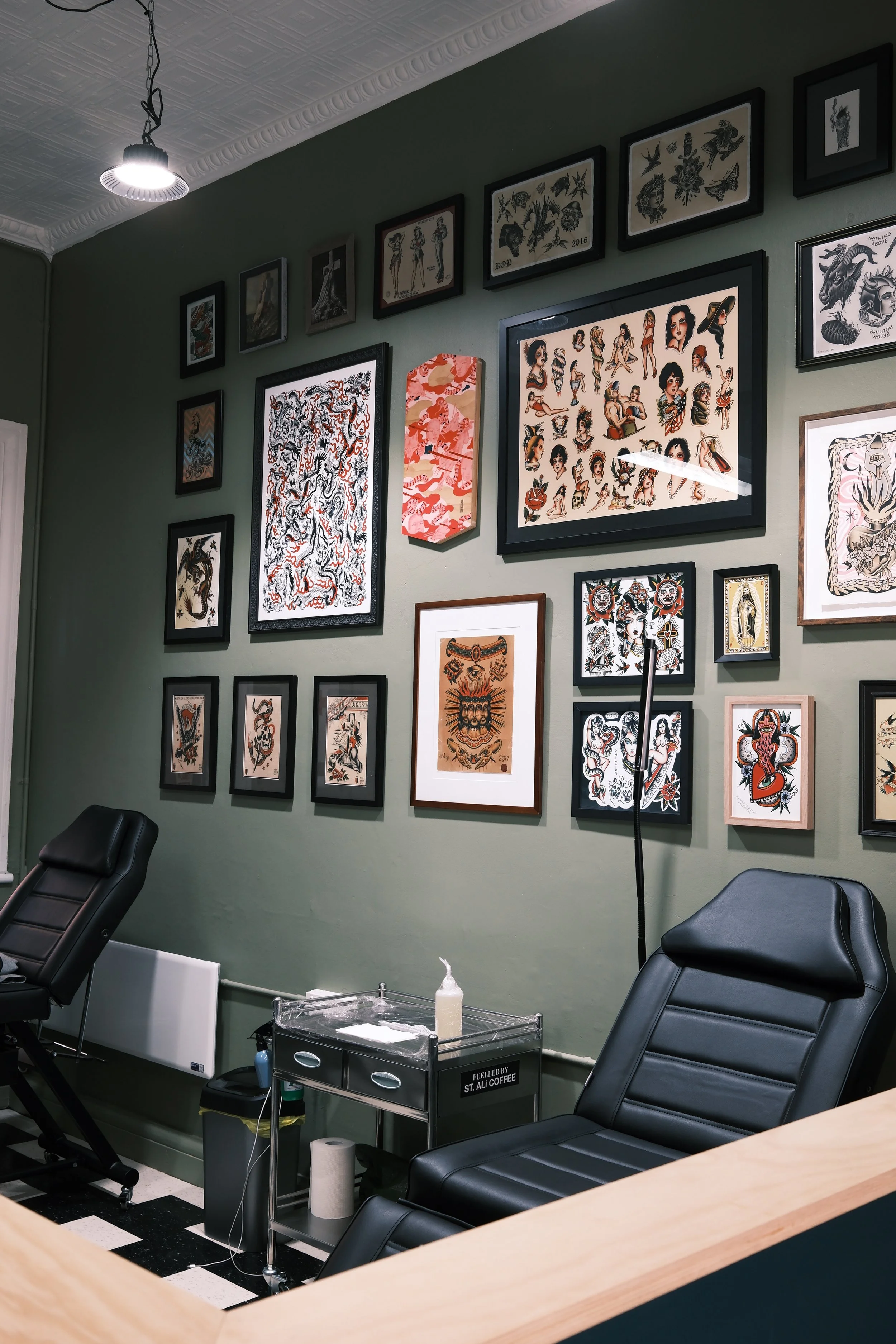 Tattoo studio with framed artwork on the green wall, black tattoo chairs, a small rolling cart with supplies, and a ceiling light.
