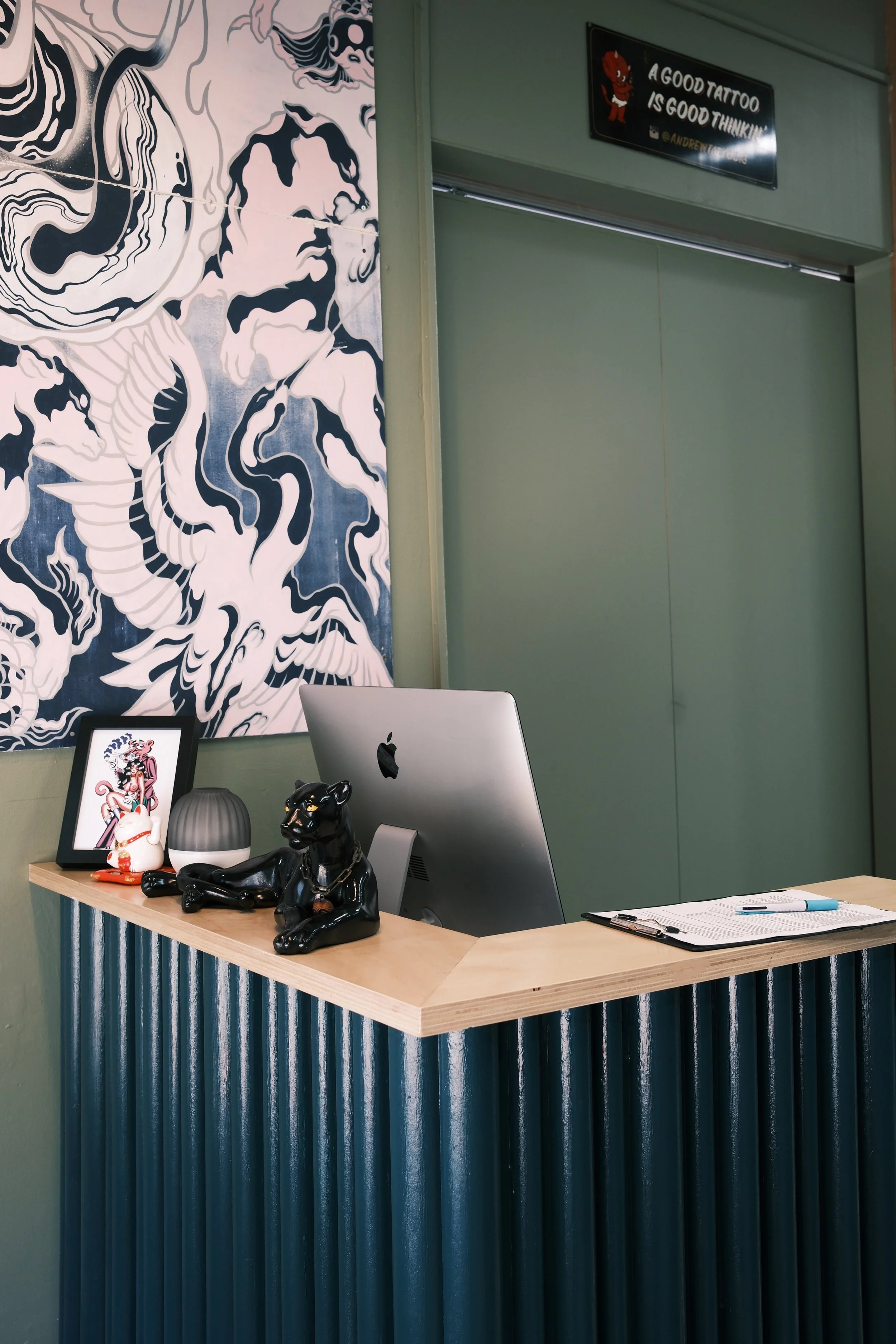 Office reception desk with decorative items, including a black panther figurine, a framed artwork, a small ceramic figurine, a diffuser, and a notebook with pens on top. Behind the desk is a large wall art with black and white abstract waves and a si