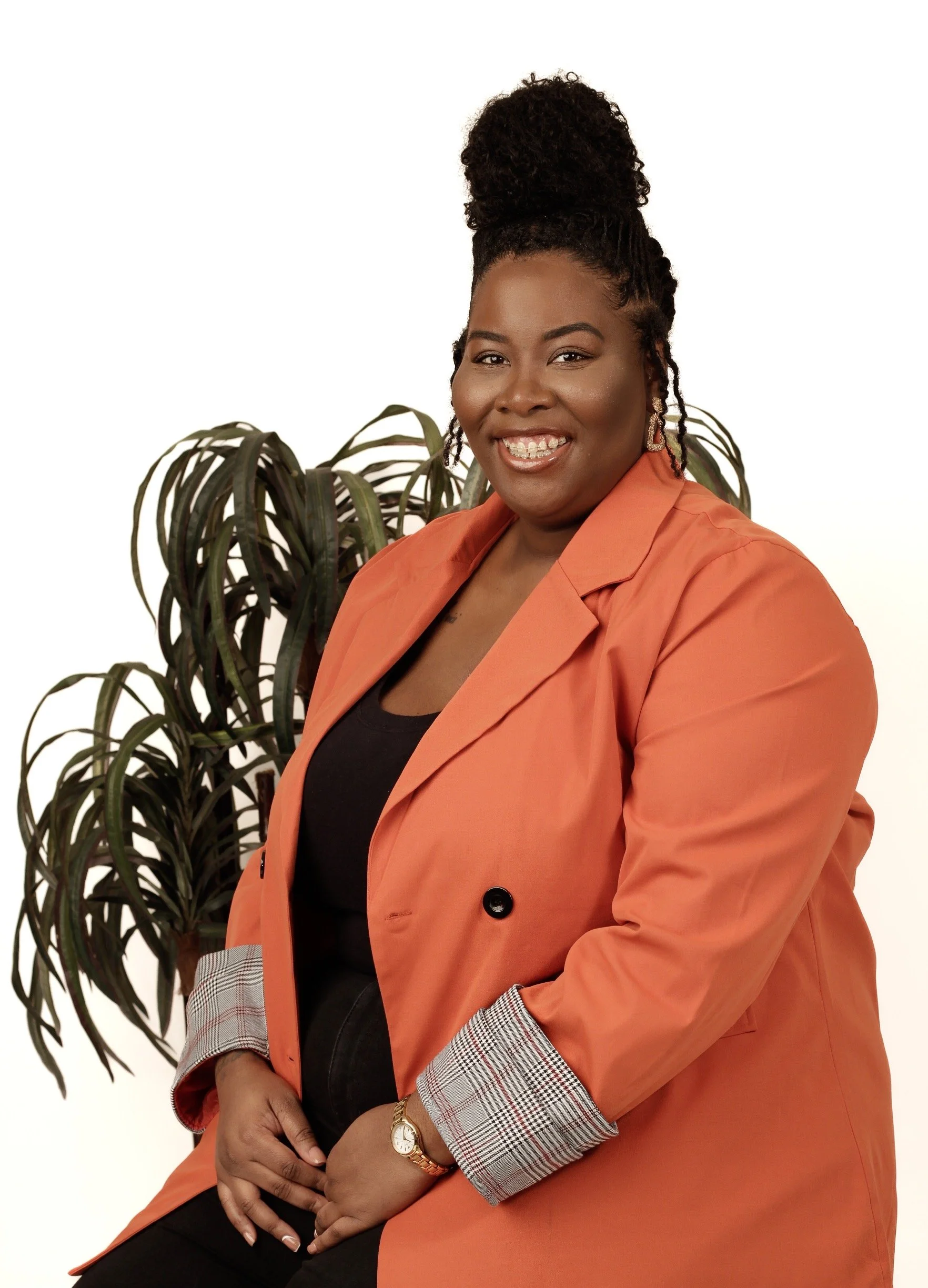 A woman with dark curly hair styled in an updo, smiling, dressed in a black top and an orange blazer with checkered cuffs, sitting in front of a large potted plant.