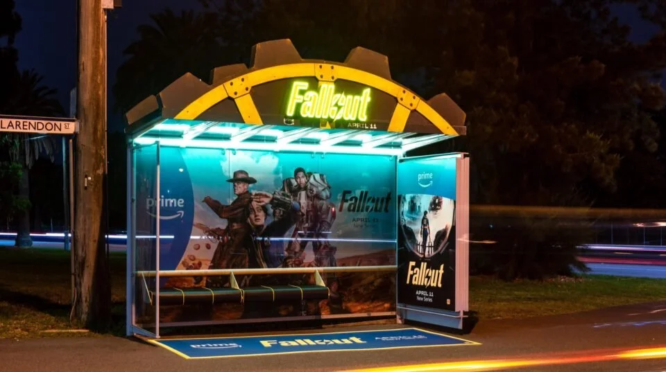 Bus stop with Fallout-themed advertisement, featuring characters from the Fallout series, illuminated at night with a neon sign overhead.