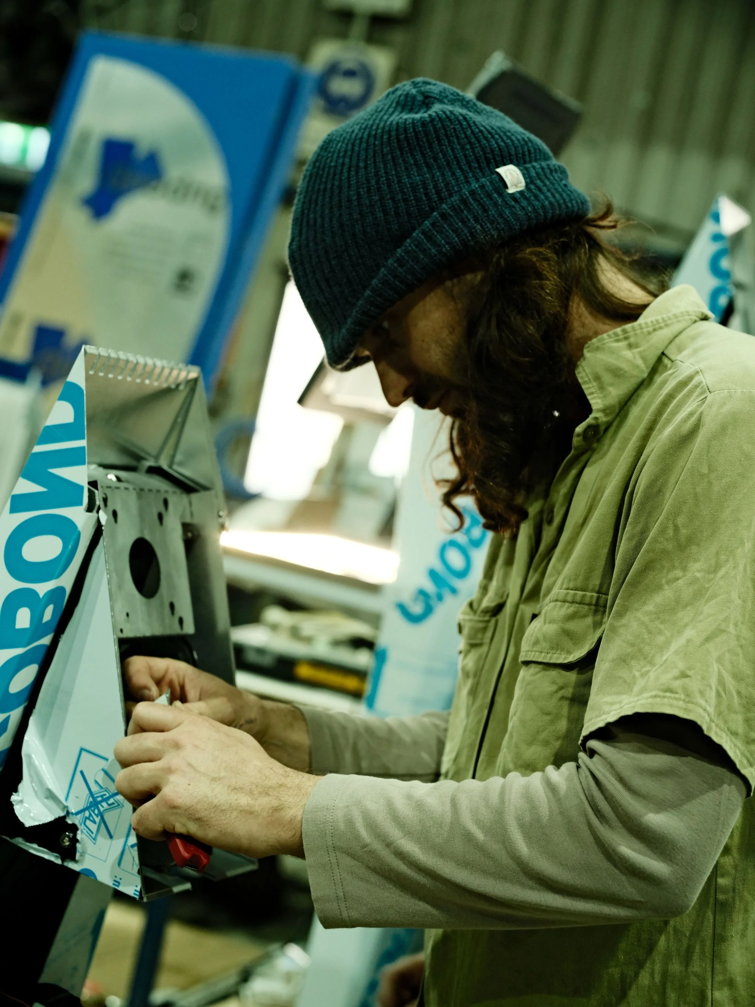 A man wearing a blue knit beanie and a beige work shirt repairs or constructs a metal object in a workshop with various equipment and signs in the background.
