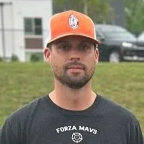 A man with a beard wearing an orange baseball cap and a black T-shirt with white text standing outdoors in a grassy area.