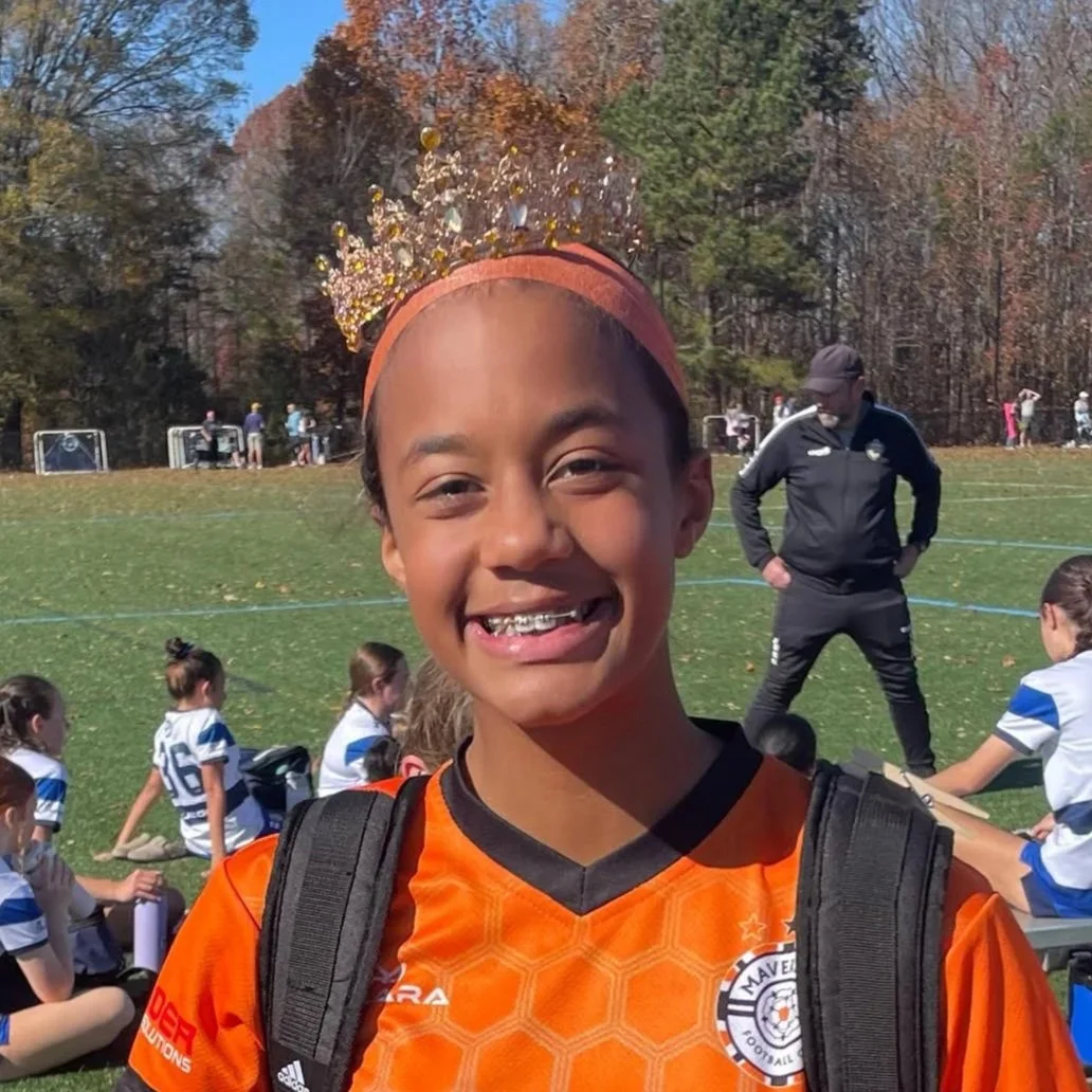 A young girl wearing an orange soccer jersey and a crown stands smiling in a grassy field, with other children in similar jerseys sitting on the ground behind her. There are trees in the background and a coach or adult figure in dark athletic wear standing behind her.