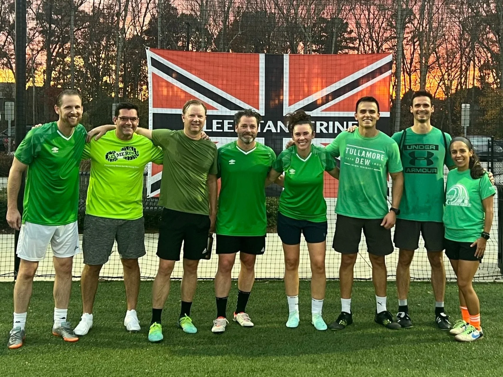Group of eight people standing on a soccer field, smiling with arms around each other, in front of a large British Union Jack flag banner during sunset.