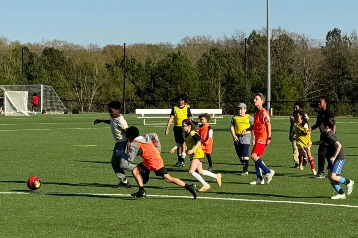 Children playing soccer on a field with a coach or referee in the background during daytime.