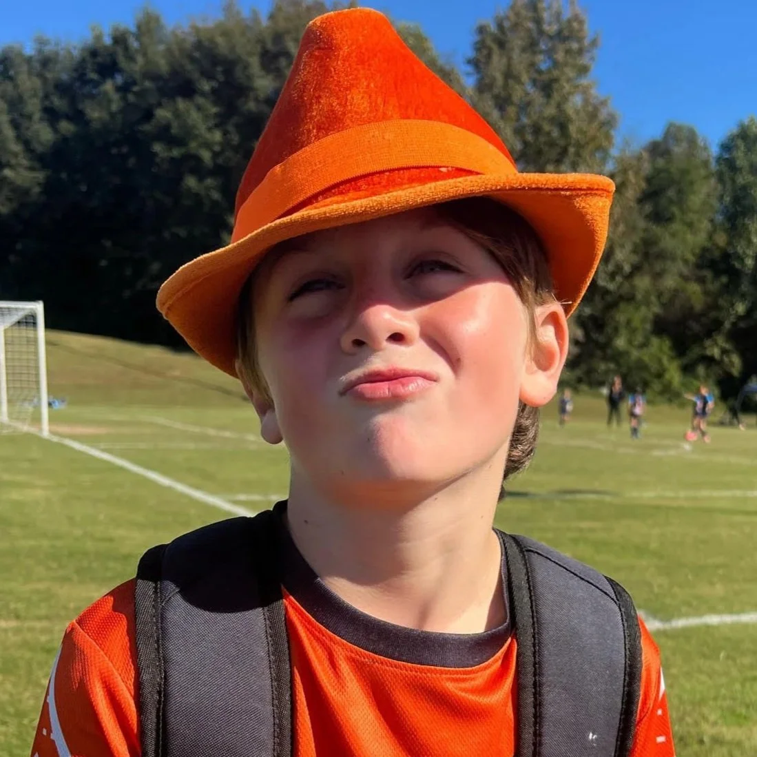 Young boy wearing an orange hat and orange sports jersey on a soccer field, with trees in the background.