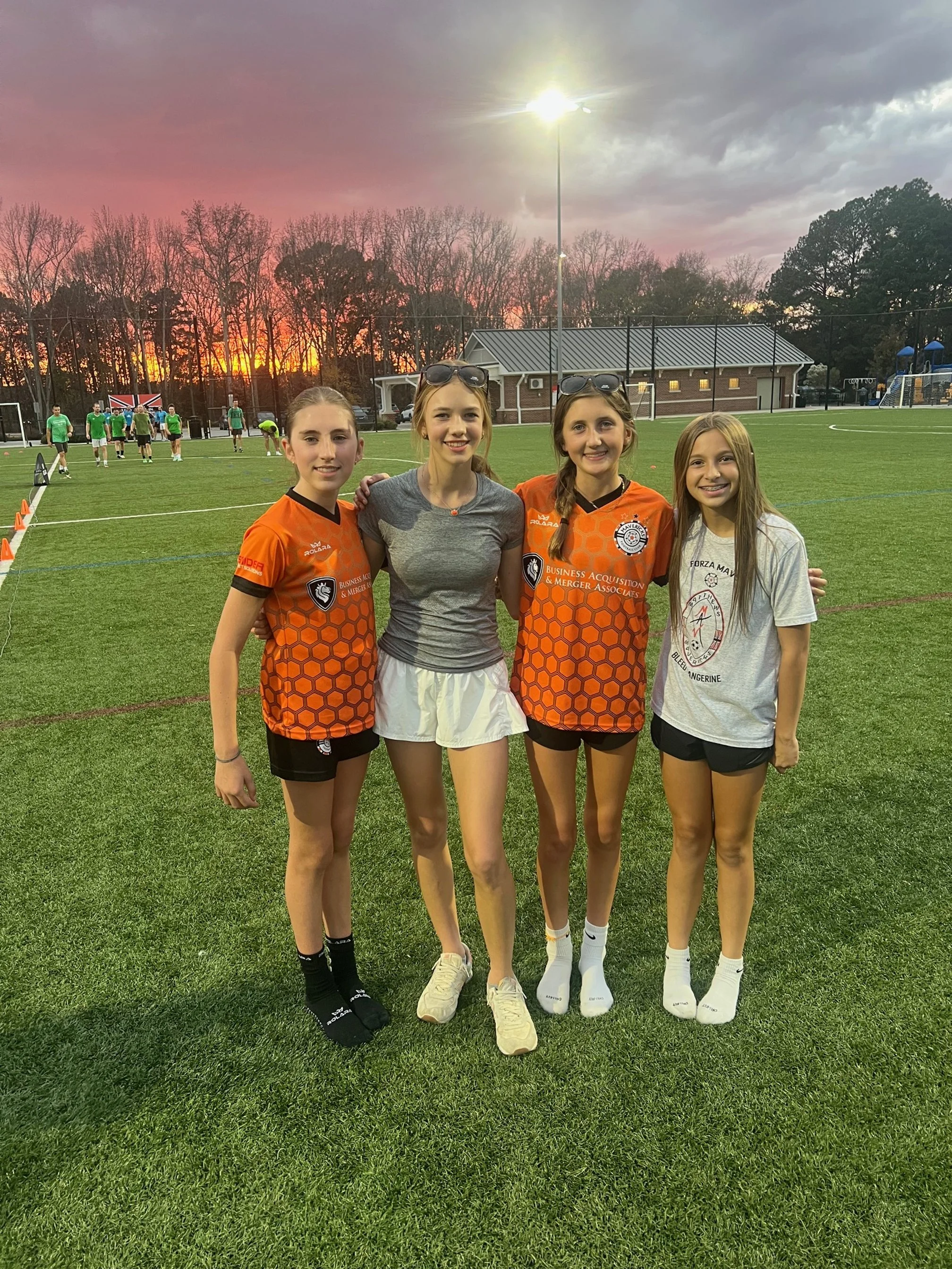 Four young women standing on a sports field during sunset, with three of them wearing orange sports jerseys and the fourth in a gray t-shirt, smiling and posing for a photo.