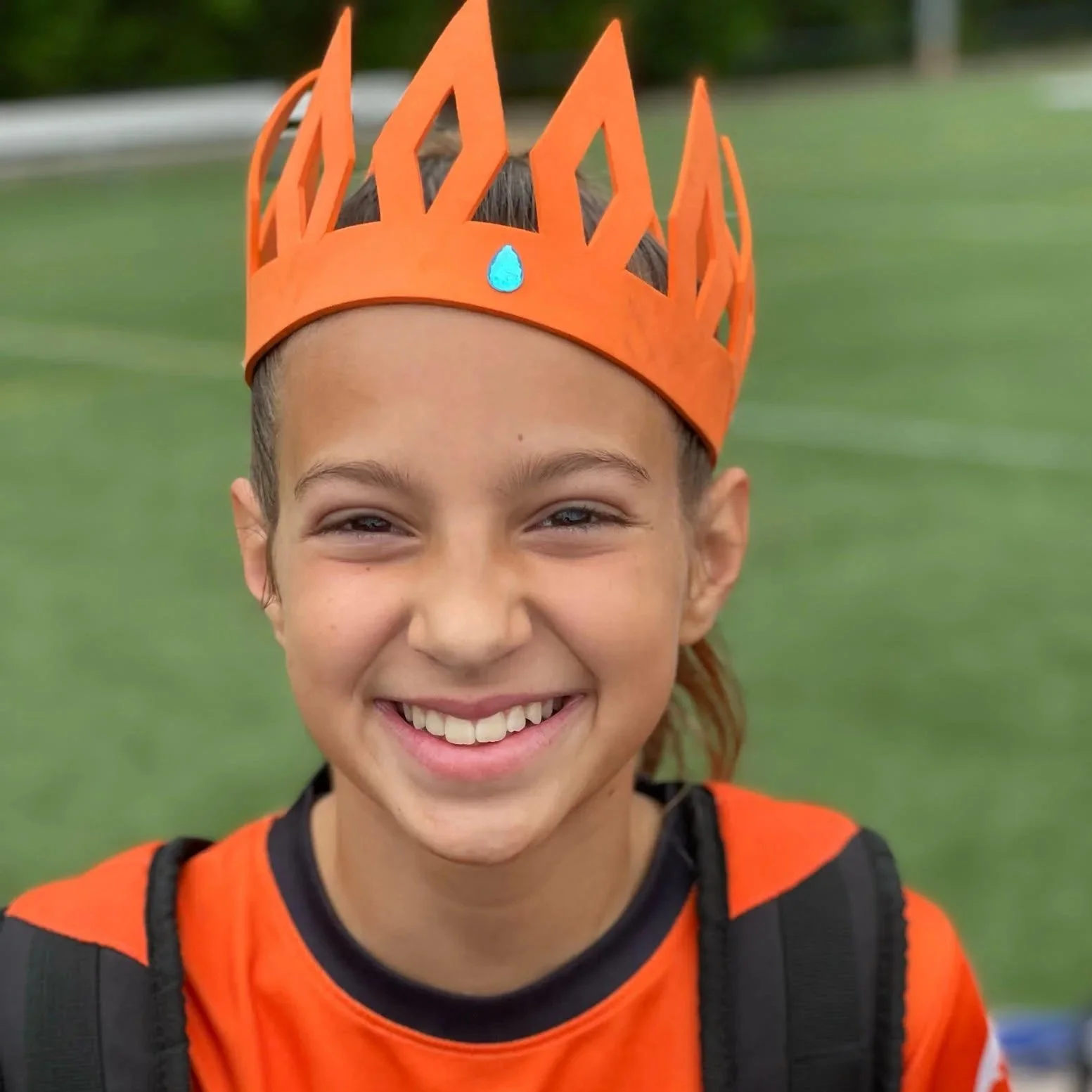 Smiling girl wearing an orange paper crown with a blue gem on a green field.