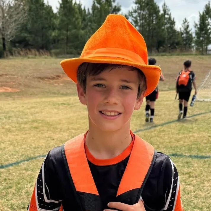 Young boy wearing an orange hat and sports uniform outdoors with other children in background