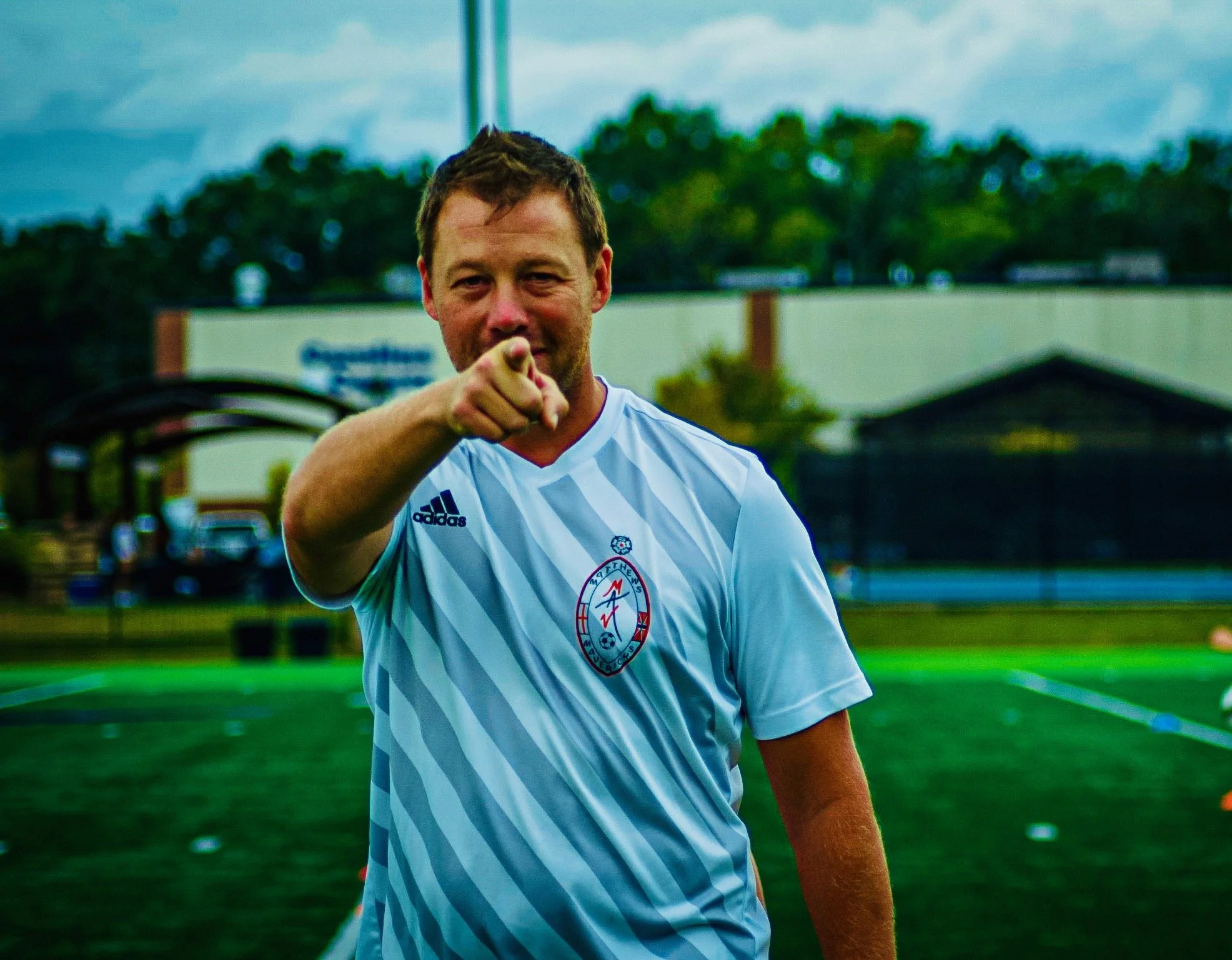 A man in a white soccer jersey pointing at the camera during a practice on a sports field.