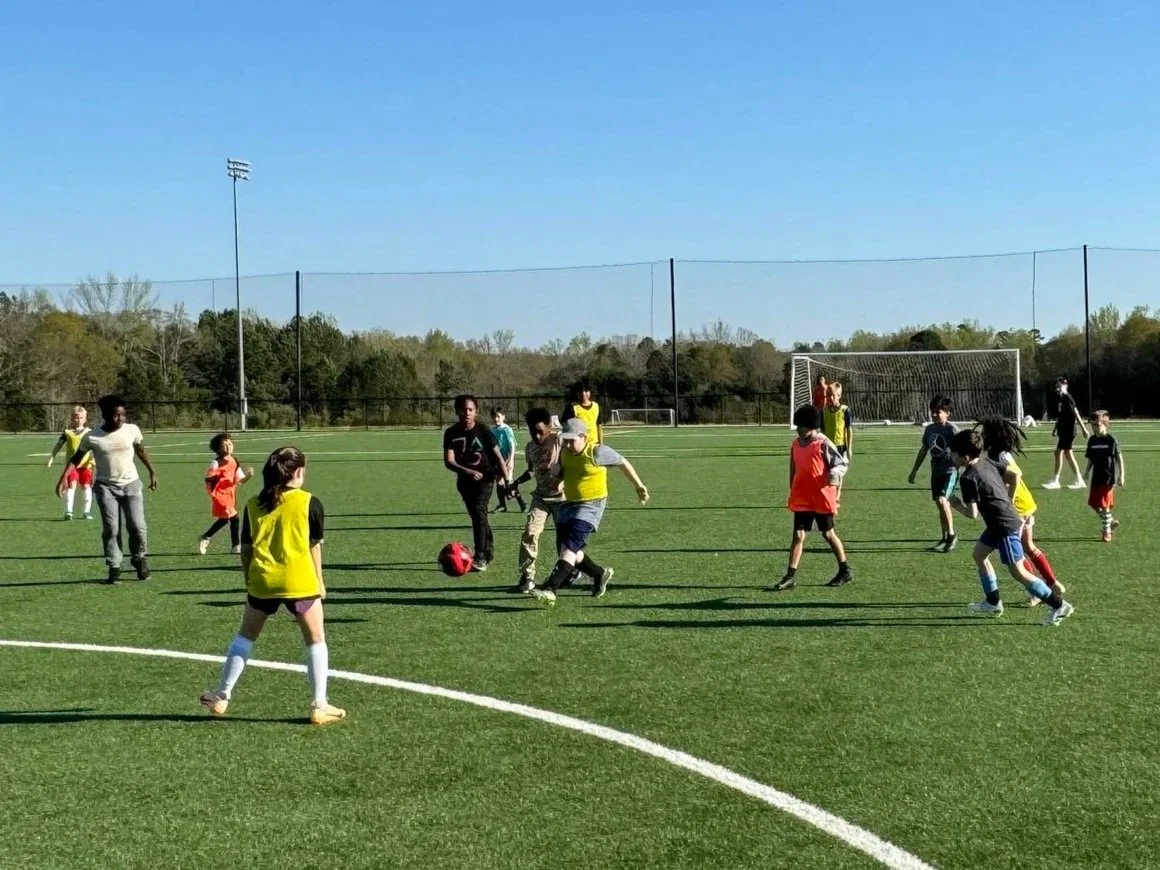 Children and adults playing soccer on an outdoor field, some wearing bright vests, under a clear blue sky.