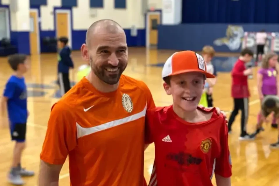 A man and a boy are smiling in a gymnasium. The man is wearing an orange sports shirt and the boy is wearing a red Manchester United jersey and a red and white cap. Several children are in the background.