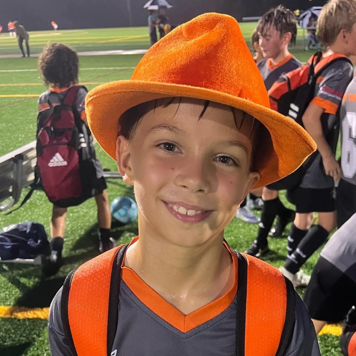 Smiling young boy wearing an orange hat and sports uniform on a soccer field, with other children and people in the background.