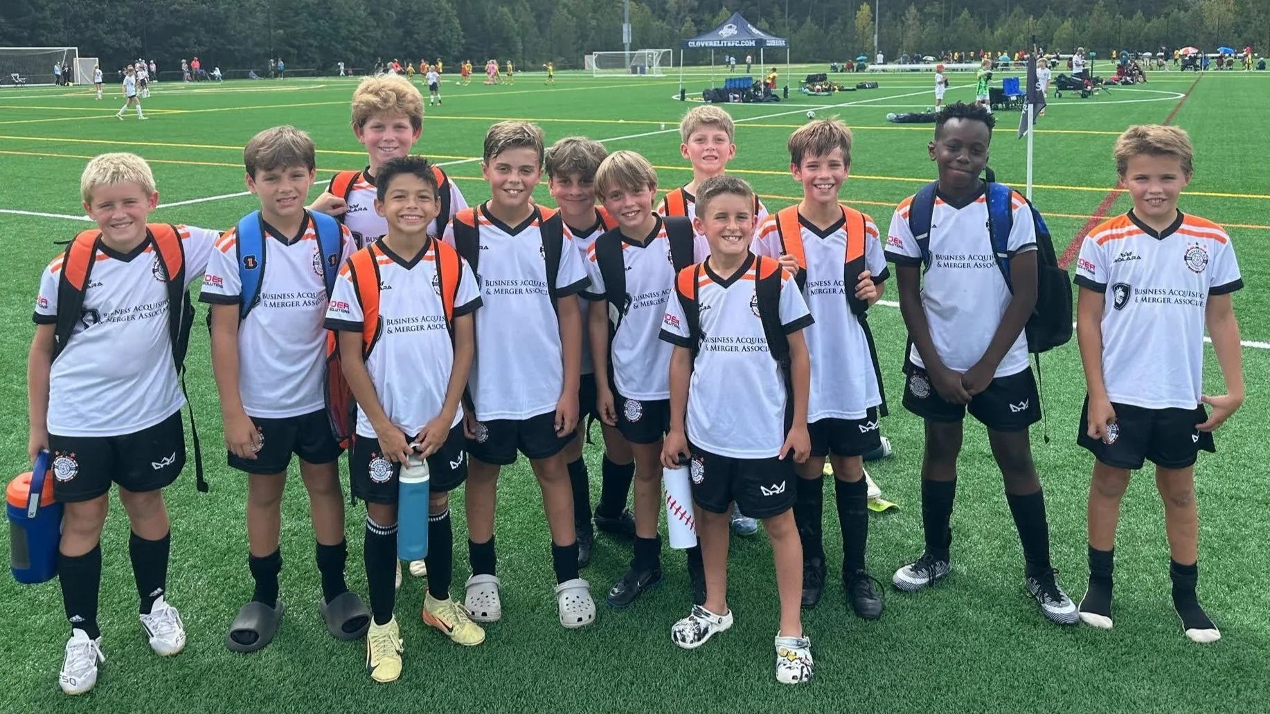 Group of young boys in soccer uniforms standing on a soccer field, smiling at the camera. They are wearing white jerseys with black shorts and black socks, some holding water bottles, with backpacks on their shoulders. In the background, there are other kids playing soccer and equipment on the field.