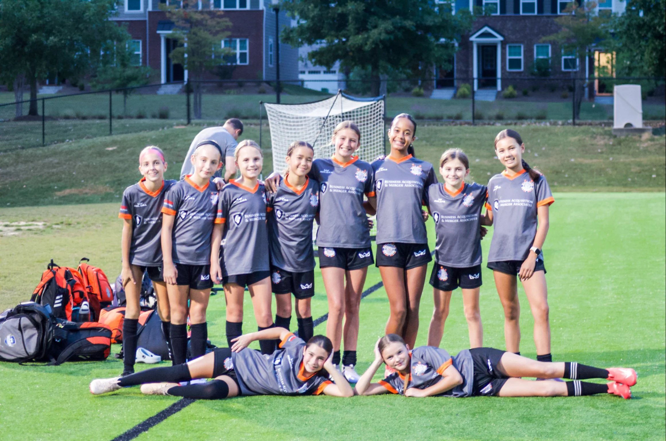 A girls' soccer team posing together on the field after a game, wearing matching uniforms, with a soccer goal in the background.
