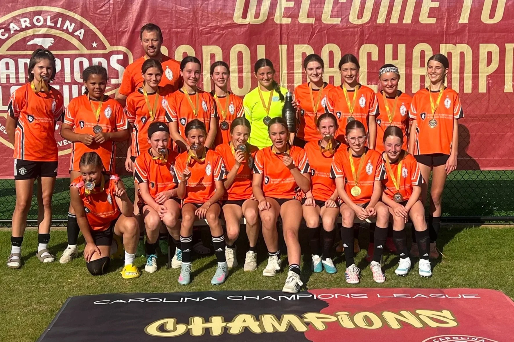 Youth girls soccer team in orange uniforms celebrating with medals and a trophy after winning a championship, with a large banner in the background.