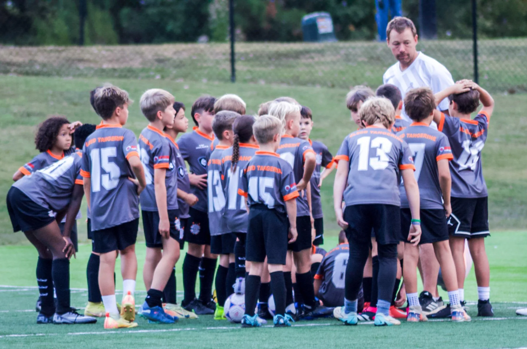 Young soccer team gathered around their coach on the field during a game or practice, with some players standing and others sitting, all wearing gray uniforms with orange accents and numbers, on a grassy field with a fence and trees in the background.