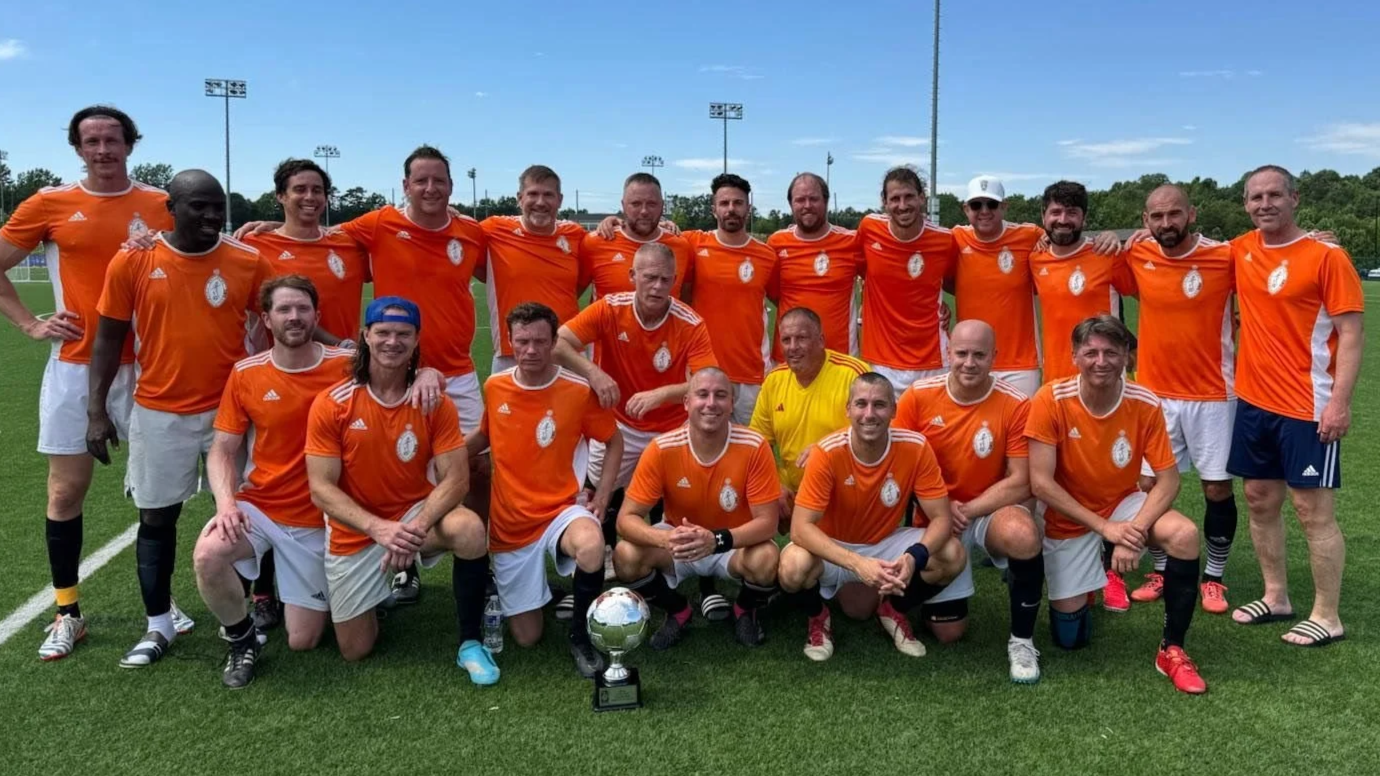 A group of men in orange soccer jerseys and white shorts, standing on a soccer field, posing for a photo with a trophy.